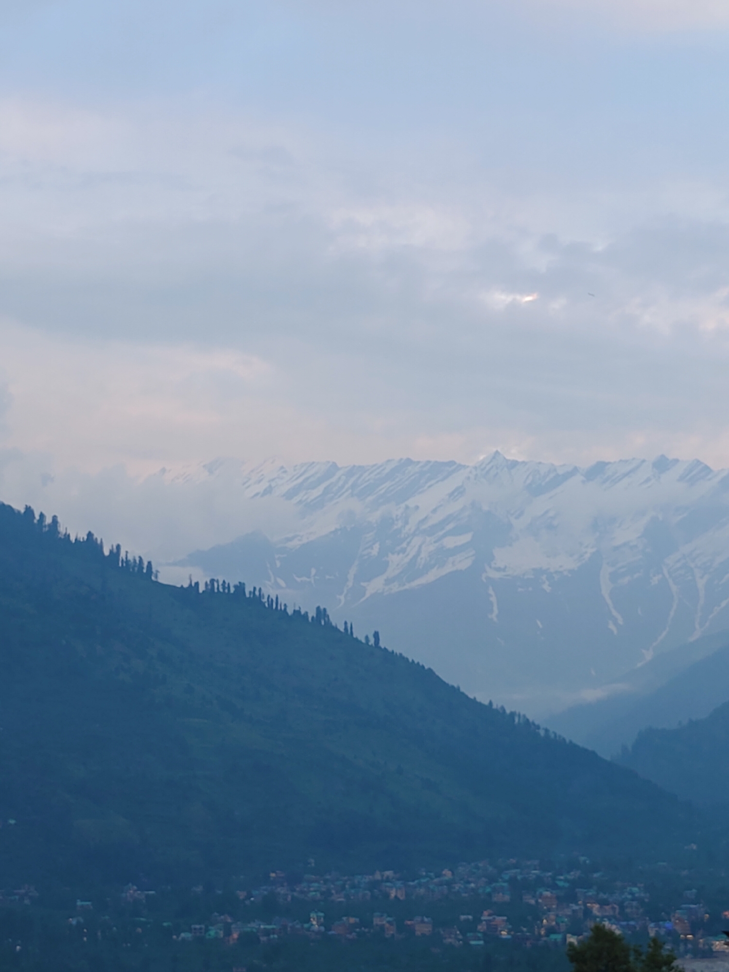 The image depicts a panoramic view of a mountainous landscape near Vashisht, India. In the foreground, a dark, densely forested mountainside slopes downwards towards a small town or village nestled in the valley. The town's lights are visible, suggesting it is either dusk or dawn.
Dominating the background are majestic snow-capped mountains, their peaks partially obscured by soft, hazy clouds. The sky above is a pale blue, with patches of clouds, some of which are illuminated by faint sunlight, hinting at either sunrise or sunset. The overall atmosphere is serene and tranquil, conveying a sense of peace and natural beauty.
The image captures a quiet moment in a mountainous region, emphasizing the grandeur of nature and the presence of human settlement within it. There is no visible text in the image.