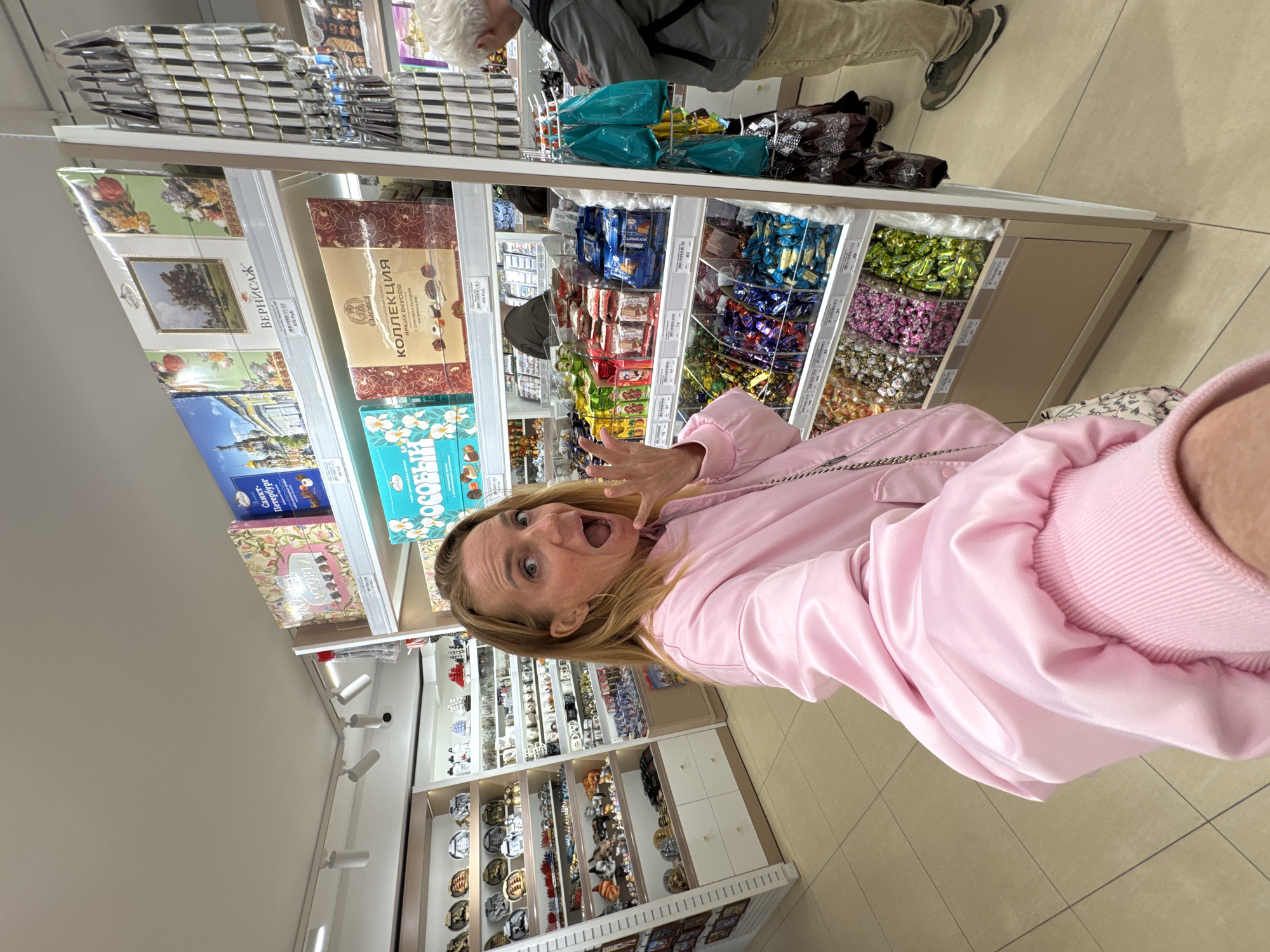 The image shows a woman in a pink jacket standing in a store, possibly in Saint Petersburg, Russia, based on the signage in Cyrillic script. She is facing the camera with an excited expression, mouth open, and hands raised near her face. Behind her are shelves filled with various packaged goods, including candies and boxed chocolates. There are also shelves displaying souvenir plates and other trinkets. Other shoppers can be seen in the background. The store has bright, artificial lighting and tiled floors.