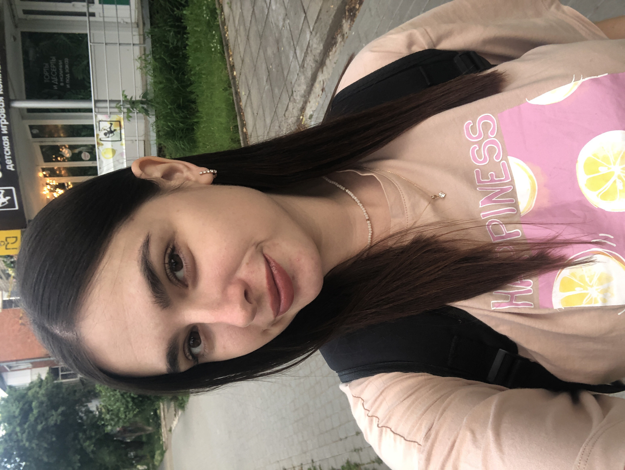 The media file is a selfie of a young woman with long, dark hair and brown eyes. She is wearing a light pink t-shirt with a graphic of lemons and the word "HAPPINESS" in bold pink letters. She is also wearing a delicate pearl necklace and a ring on her left hand.
The background suggests an outdoor setting, likely a street or sidewalk, with a building that appears to be a shop or cafe. There is greenery visible, possibly a grassy area or trees. The building has large windows and signage. Some visible text on the building's signage appears to be in Russian, with phrases like "ДЕТСКАЯ ИГРОВАЯ КОМНАТА" (Children's Playroom) and "ТОРТЫ И ДЕСЕРТЫ" (Cakes and Desserts).
The woman is looking directly at the camera with a slight smile. Her expression appears relaxed and content. The lighting suggests it is daytime, but the exact time of day and weather conditions are not clearly discernible. The orientation of the photo is slightly tilted. The context of Chaikovsky, Russia is provided.