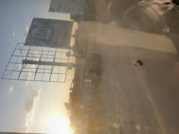 A low-angle shot captures a partly cloudy sky at sunset. Silhouetted billboards stand tall, their metal frames visible. A hazy atmosphere obscures details of the background. The scene seems to be viewed through a grimy window, distorting the light and creating a smeared effect. The sun casts a warm glow, emphasizing the billboards' forms.