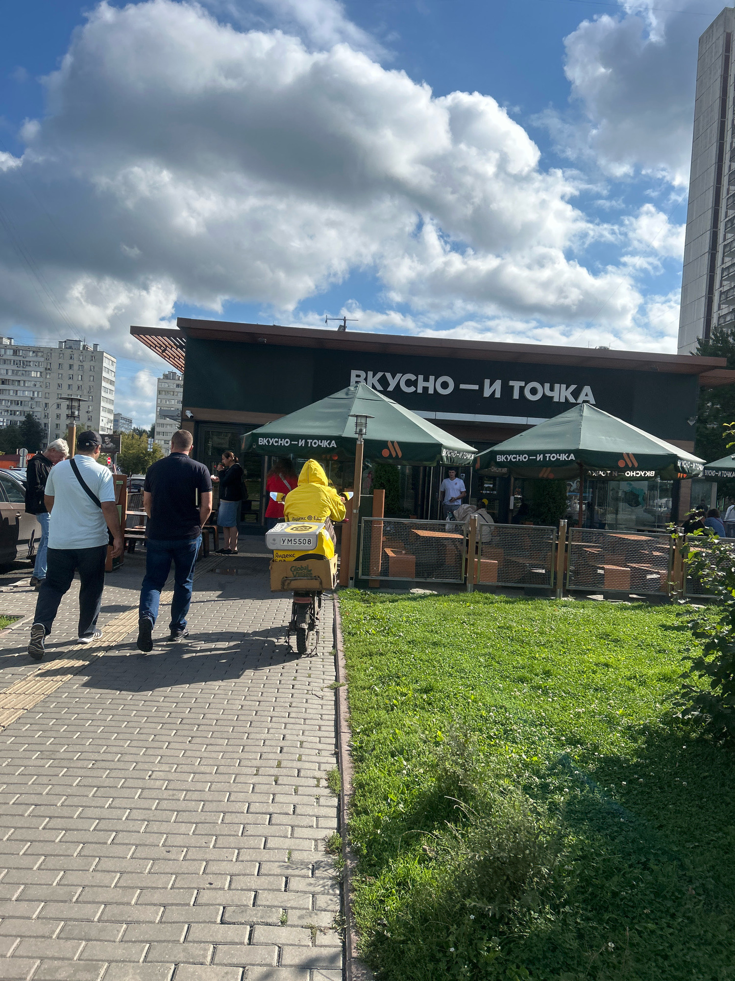 The image depicts the exterior of a "Vkusno - i tochka" (Russian for "Tasty - and that's it") fast-food restaurant, formerly a McDonald's, located in Moscow, Russia. In the foreground, two men are walking away from the camera on a paved walkway. To the right of the walkway, a grassy area is visible, bordering a section of the restaurant. A person in a bright yellow hooded jacket is riding a bicycle with a delivery box on the back, moving in the same direction as the men. The bicycle's license plate reads "YM5508." The restaurant building features a dark facade with the "Vkusno - i tochka" logo. Several green umbrellas, also bearing the restaurant's branding, are set up outside. Behind the restaurant, a tall, modern apartment building is visible on the left, and another taller, light-colored building is on the right. The sky is partly cloudy with large, white cumulus clouds against a blue background. The overall scene suggests a typical urban environment on a sunny day, with activity around a popular fast-food establishment.