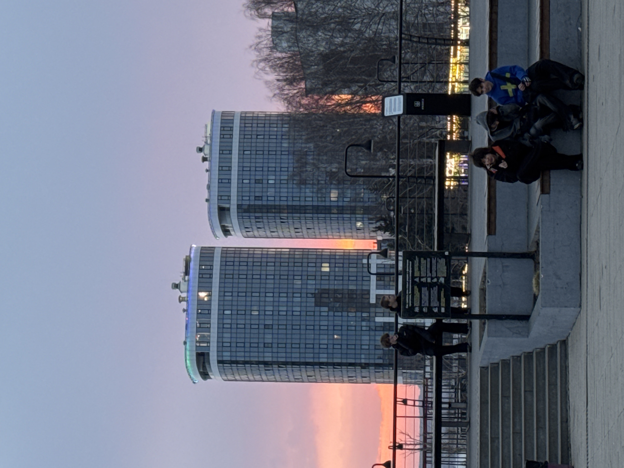 Two cylindrical residential buildings with glass facades stand against a sunset sky. In the foreground, people are seen sitting and standing on a concrete structure with stairs and railings. The location is Izhevsk, Russia.