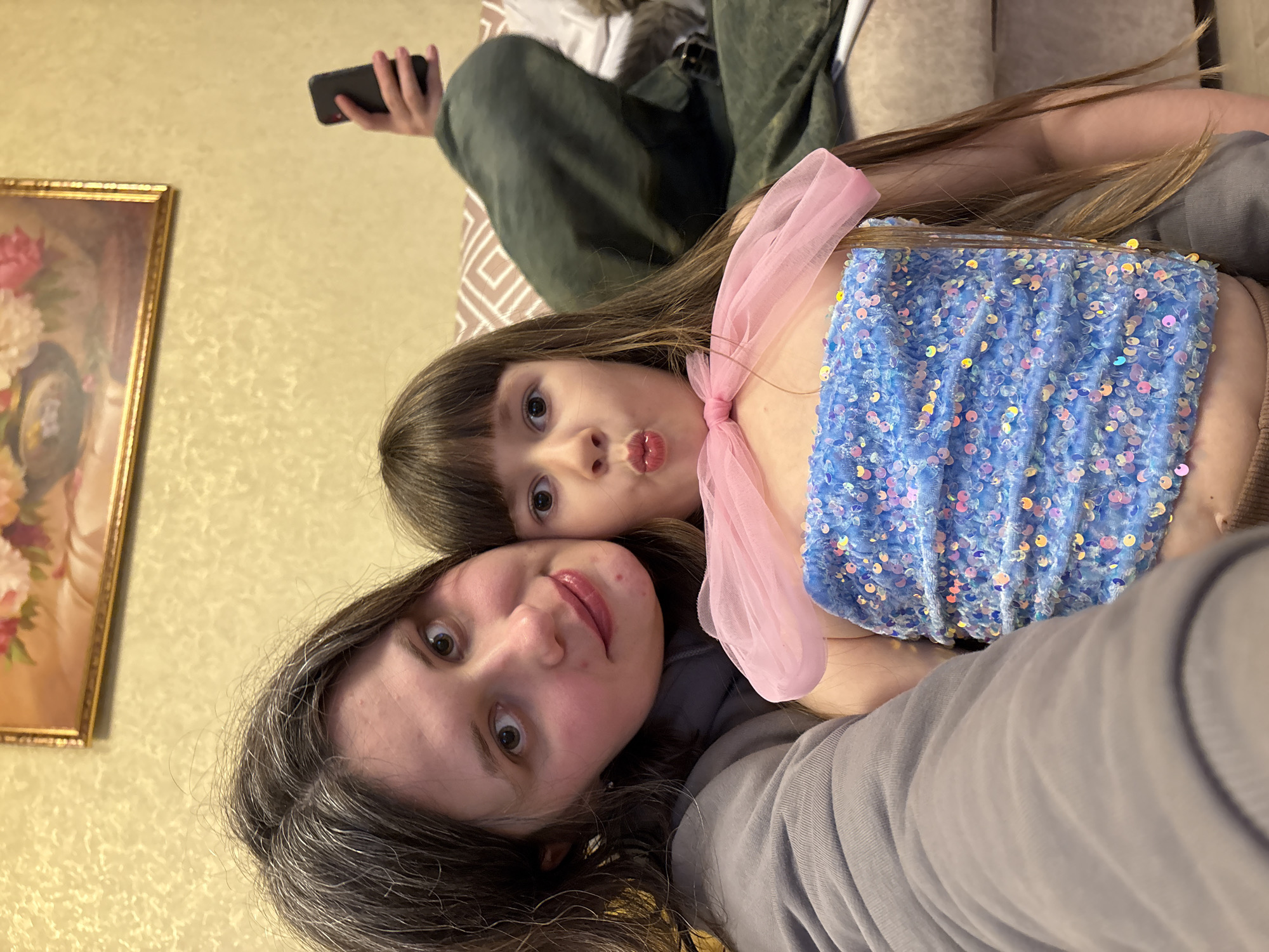 A selfie shows a woman and a young girl. The woman has long, grey hair and a grey shirt. The girl has brown hair, a blue sequined top, and a pink bow. Another person is visible holding a cell phone on a couch. A painting of flowers is on the wall. The scene appears to be indoors, possibly in a residence in Chaikovsky, Russia.