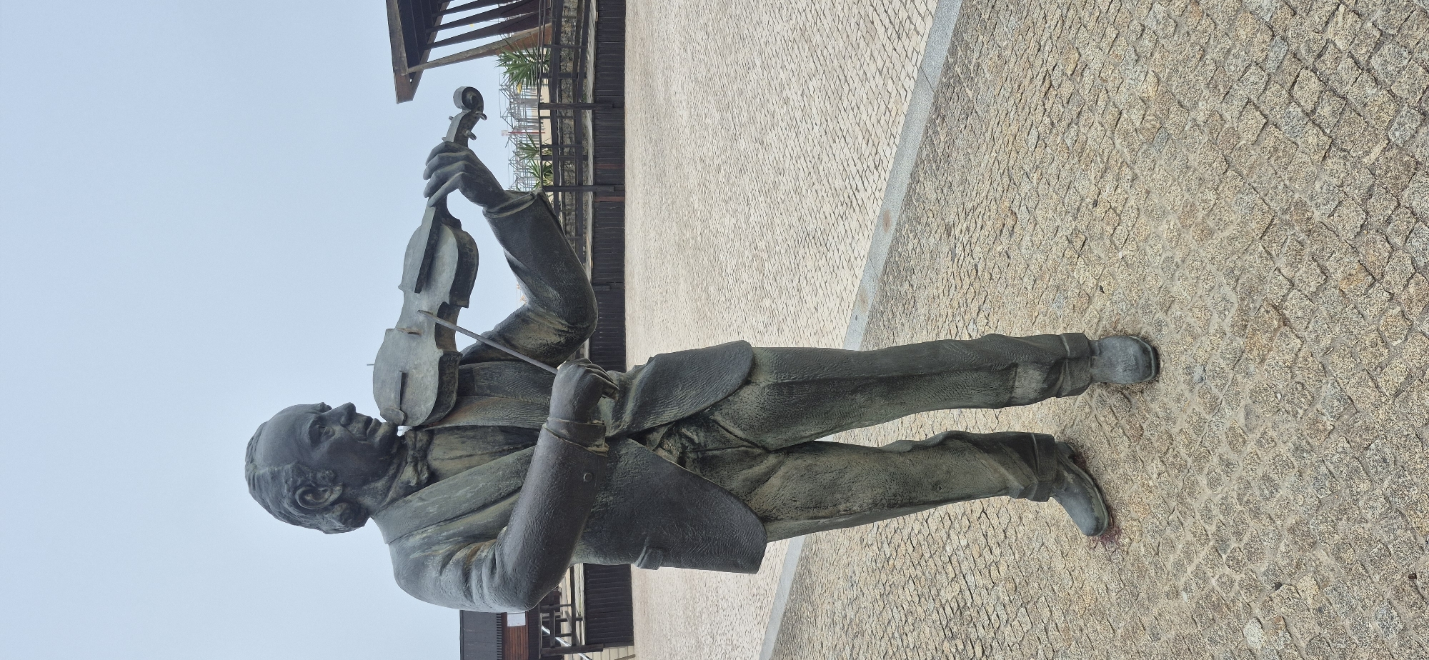 A bronze statue of a man playing a violin stands in an outdoor plaza paved with granite cobblestones. The statue depicts a man in a suit and bow tie, holding the violin with his left hand and the bow with his right. He is looking to his left. In the background, there is a wooden structure and a fence. The sky is overcast. This is a statue of composer and violinist Francisco de Lacerda located in the Praça da Lacerda, Póvoa de Varzim, Portugal.
