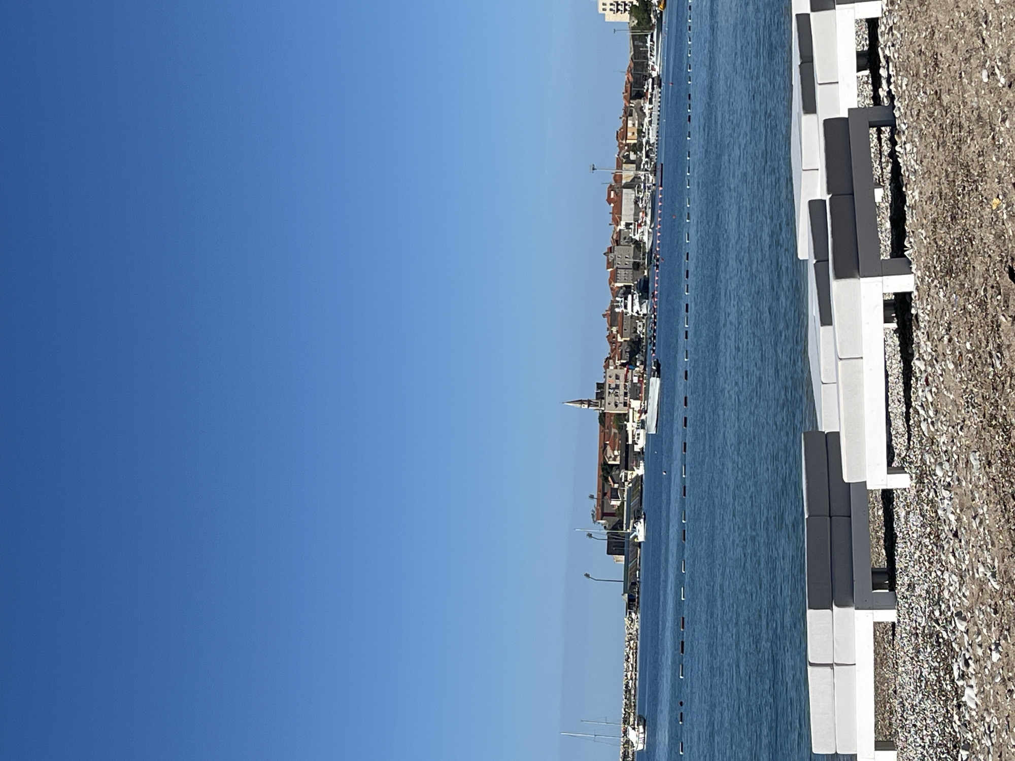 The image shows a daytime scene in Budva, Montenegro, on a clear day with a bright blue sky. The view appears to be taken from a low vantage point near the shoreline. In the foreground, there are white and gray concrete structures which appear to be part of a seawall or pier, alongside a pebble beach. The sea is visible, extending toward the horizon, with a line of buoys marking a boundary in the water. Across the water, a line of buildings and structures is visible, representing the urban landscape of Budva. There is a prominent church steeple among the buildings. In the far distance, another taller building can be seen. The image is oriented sideways. There are no people visible.