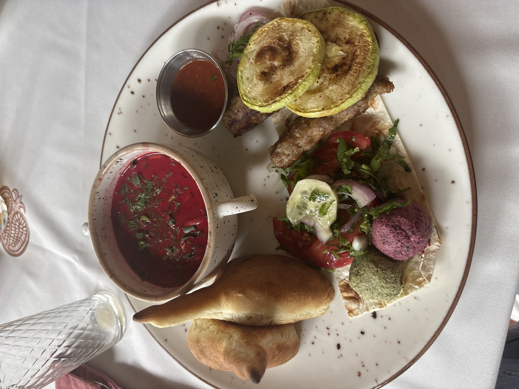 The image shows a plate of food on a white tablecloth, likely in a restaurant setting in Sochi, Russia. The dish features several items: a bowl of vibrant red soup, possibly borscht, garnished with fresh herbs and green onions; two pieces of bread; a skewer of grilled meat, likely kebabs, served with grilled zucchini slices and red onion; a small metal dish of red sauce; and a wrap filled with salad, including tomatoes, cucumbers, red onion, and cilantro. There are also two distinct balls of dip on the wrap, one purple and one green. A patterned glass bottle is visible to the left of the plate, and a part of a decorative pineapple motif is also visible on the tablecloth. The lighting suggests it is daytime. There is no visible text in the image. The overall impression is a hearty and diverse meal.