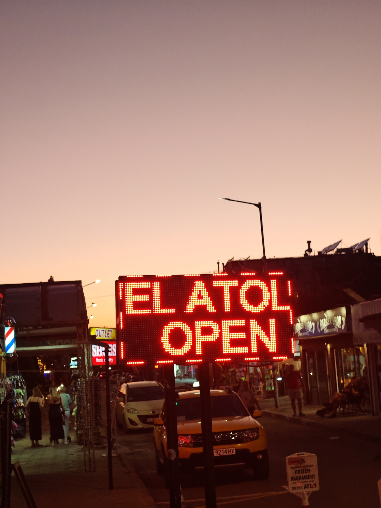 The image shows a street scene in Nesebar, Bulgaria at dusk. The sky is a gradient of orange and pink. In the foreground, an LED sign displays "EL ATOL OPEN" in bright red lettering. Below the sign, a yellow car with the license plate "9214HX" is parked.
To the left, people are walking past shops, including one with a barber pole and another with a sign that reads "OUTLET SH: 38 Best&ity". Across the street, there is a business identified as "BARBER SHOP". A small white sign on the curb has text written in Bulgarian "ПЛАТЕН АБОНАМЕНТ ХОТЕЛ АТОЛ". A lamppost is visible in the background.
