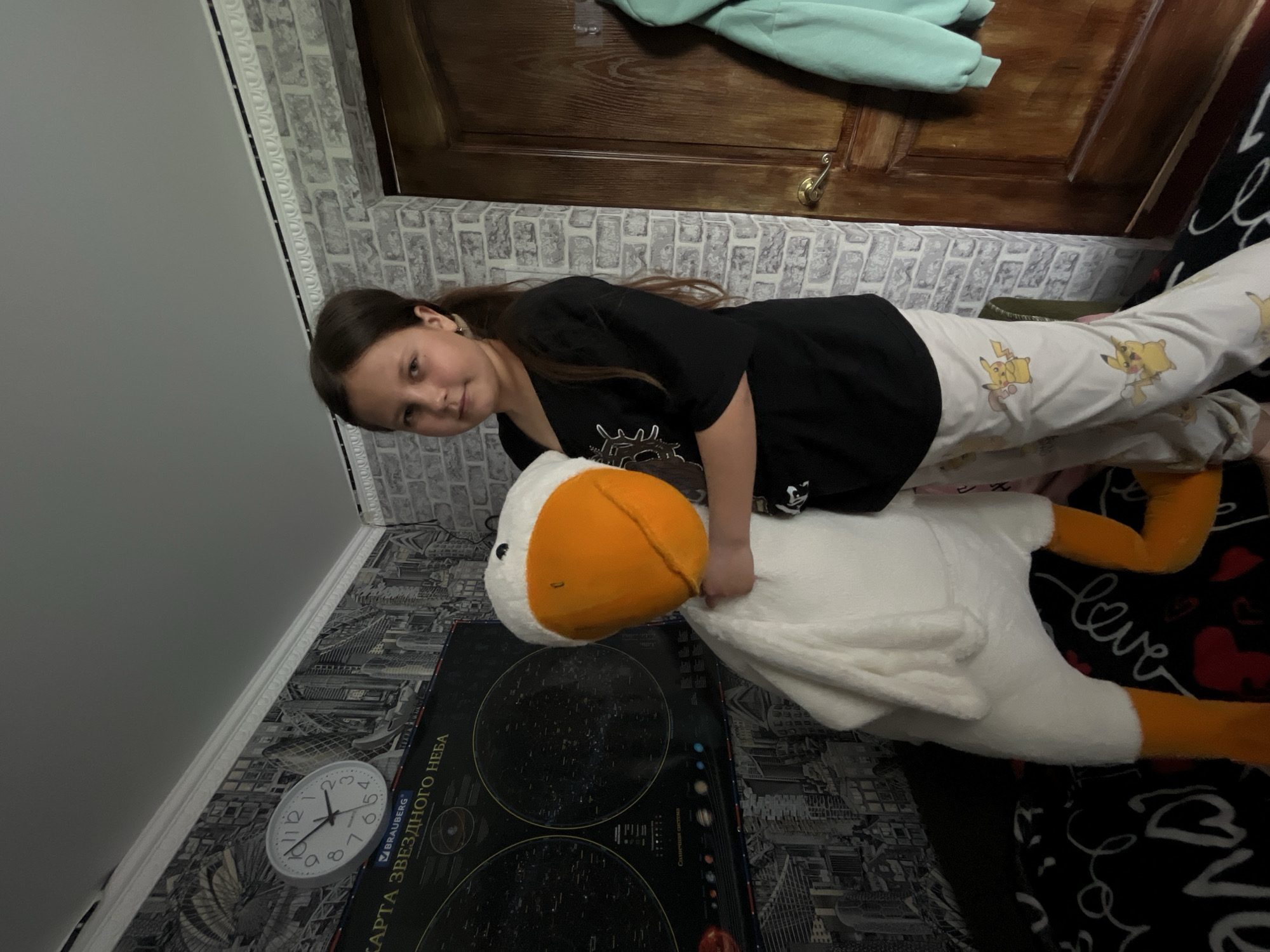 A girl is lying on top of a large plush goose, resting her torso on its back and gripping its neck. She has long brown hair and is wearing a black t-shirt and light-colored pajamas patterned with Pikachu characters.
The goose is mostly white with an orange beak and legs. Beneath the goose is a rectangular poster or placemat featuring star charts and text in Russian, including "КАРТА ЗВЕЗДНОГО НЕБА" (Star Chart).
The background includes patterned wallpaper with a brick-like design, a wooden door, and a clock hanging on the wall. The overall setting appears to be indoors, possibly a bedroom.
While the location is specified as Novopokrovskaya, Russia, there is no discernible information in the image that confirms this. Therefore, that detail cannot be factually determined from visual evidence.
