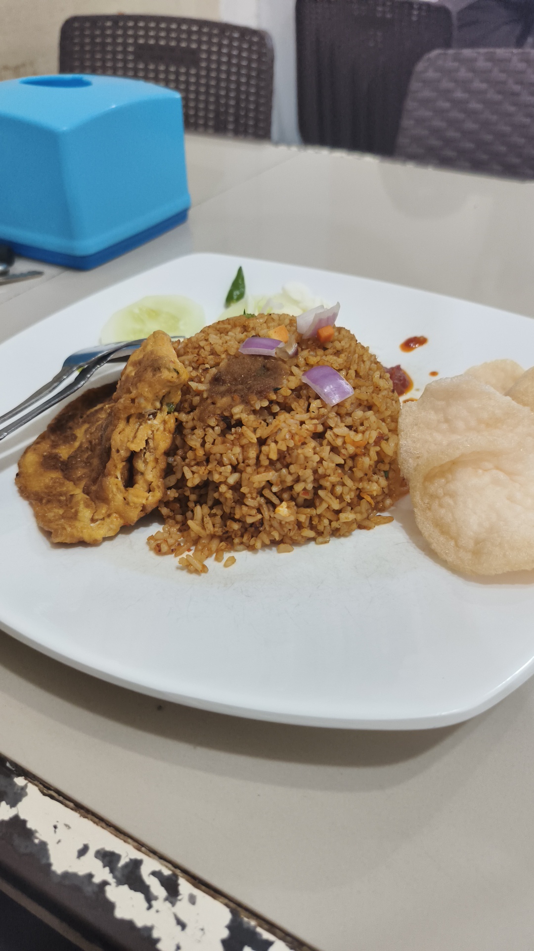 A close-up shot shows a plate of fried rice with omelet and crackers, presented on a white square plate. The fried rice is a rich brown color, mixed with small pieces of vegetables and topped with a small piece of meat or protein, and garnished with thinly sliced red onion and a green chili pepper. To the left of the rice is a folded omelet. On the right side of the plate are several light-colored, puffed crackers. A fork and knife are visible to the left of the omelet. The plate sits on a beige surface, with a blurred background showing a blue tissue box and dark chairs, suggesting a dining setting, possibly a restaurant. The lighting is somewhat dim, indicating it could be evening or indoors. The overall scene depicts a prepared meal, ready to be eaten, with a focus on the food. No text is visible in the image. The location context of Lhokseumawe, Indonesia, suggests this is likely a local Indonesian dish.