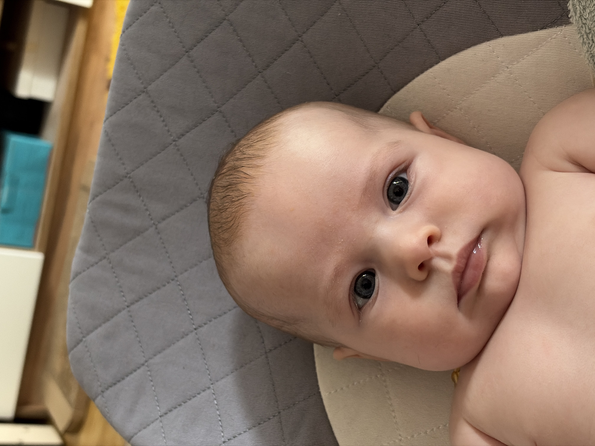 A baby lies in a padded seat. The seat has a gray quilted fabric on the left and a beige fabric on the right. The baby has fair skin, light hair, and blue eyes. The baby's face is facing the viewer. The background shows part of a wooden cabinet and a blue object. The location is unknown and there is no indication that the image was taken in Chaikovsky, Russia.