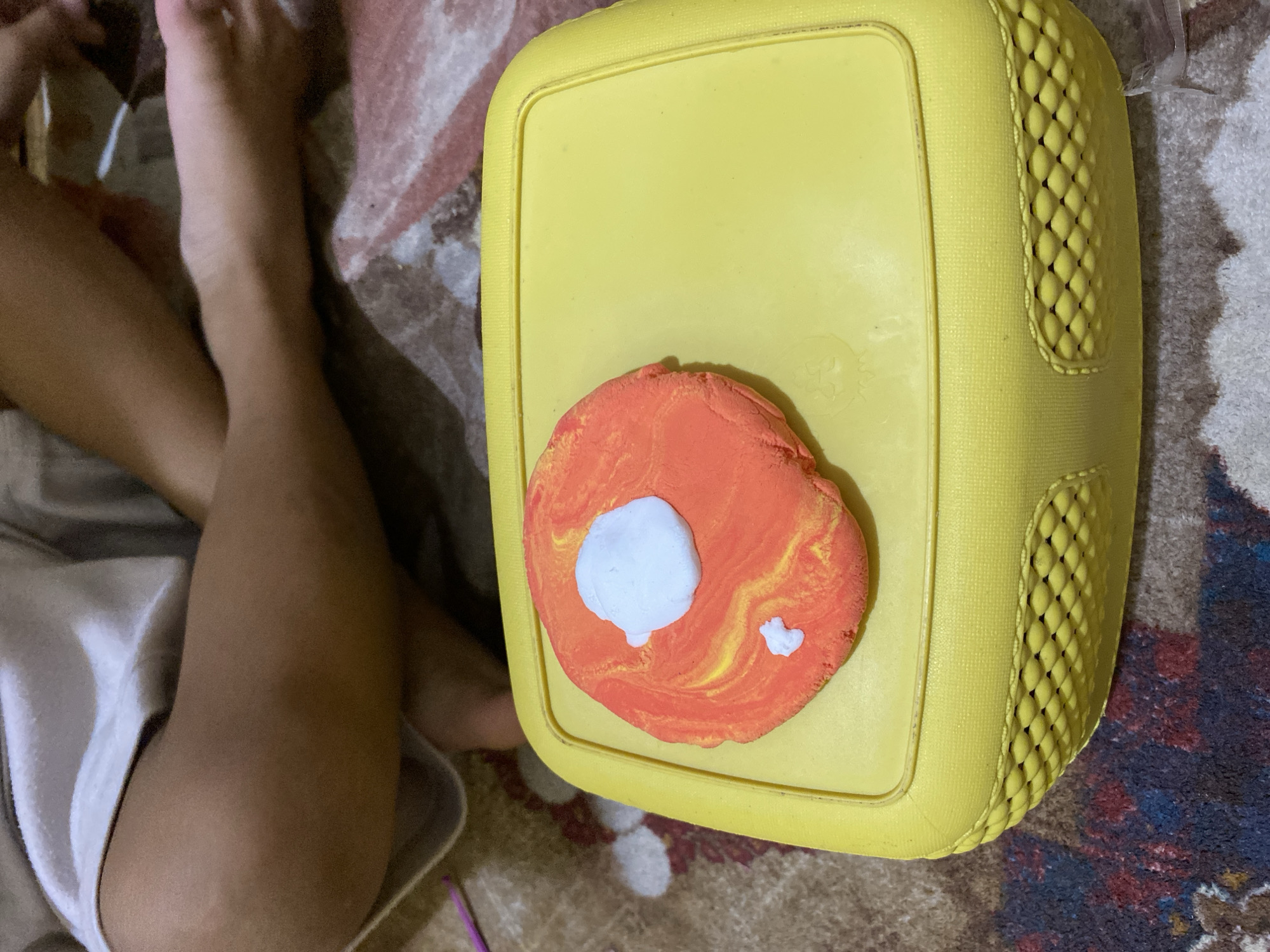 In the city of Lambaro Kueh, Indonesia, an overhead shot captures a yellow plastic container, likely a storage box, placed on a patterned rug. The container's lid features a circular indentation where a flat, orange and yellow swirled object, possibly made of clay or Play-Doh, is situated. Two small white blobs are affixed to this orange object. To the right of the container, the legs of a person, appearing to be a child due to their size and smooth skin, are visible, draped in a white cloth. The rug beneath has a marbled pattern of reds, browns, and blues, suggesting an indoor setting.