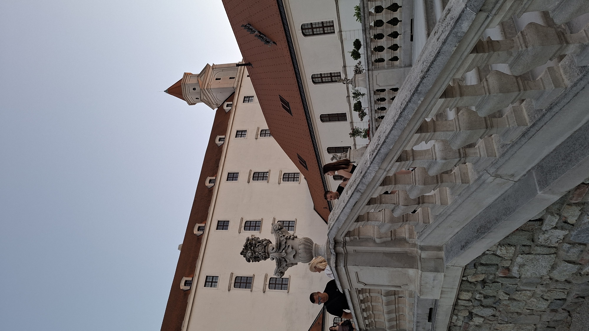 The image depicts Bratislava Castle in Bratislava, Slovakia, during what appears to be late afternoon or early evening. The castle complex is characterized by its white facade and reddish-brown tiled roofs, with a prominent tower rising in the background. In the foreground, a stone balustrade and steps lead upwards, with a decorative stone urn on the left. Several individuals are visible, some walking on the steps and others standing near the balustrade. The sky is a clear, pale blue. The overall scene conveys a sense of architectural grandeur and a leisurely atmosphere.