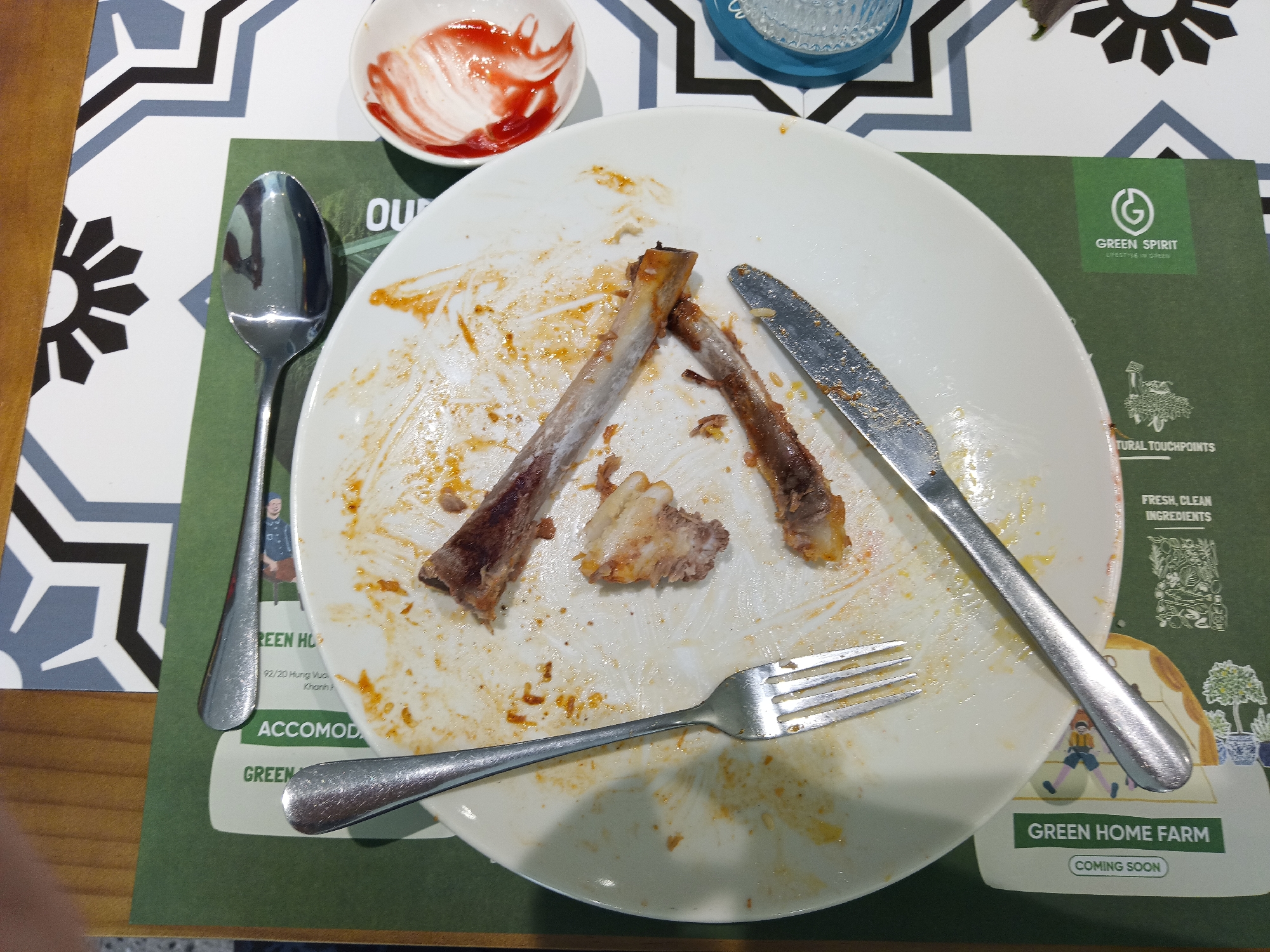 The scene depicts a dirty, finished plate of food in what appears to be a restaurant or eatery. The plate contains leftover bones, sauce residue, and other food scraps. A knife and fork are positioned across the plate, along with a spoon to the left. A small bowl with some red sauce is placed to the upper left of the plate. The plate sits on top of a green placemat advertising a "Green Spirit" and "Green Home Farm". The placemat appears to have text in English, but also suggests Vietnamese, as the text "92/20 Hung Vu" and "Khanh H." are visible, referring to a location in Nha Trang Ward, Vietnam. The table has a black and white patterned surface.