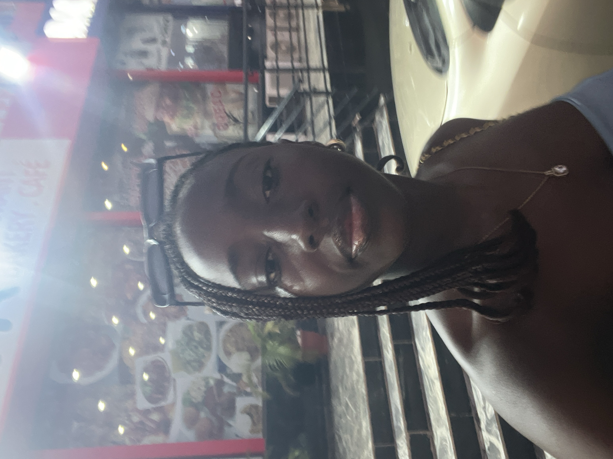 This image depicts a close-up, chest-up view of a young, dark-skinned woman at night in the city of Abuja, Nigeria. She is positioned slightly off-center to the right, looking directly at the camera with a neutral expression. She has braided hair, with some strands falling over her right shoulder, and is wearing sunglasses pushed onto the top of her head. A light-colored (possibly cream or pale blue) strapless top is visible, along with a thin gold chain featuring a small pendant around her neck and a gold hoop earring on her visible right ear. Her lips have a glossy appearance.
In the background, behind the woman's left shoulder, is the illuminated window display of a commercial establishment. The window is framed in red and showcases multiple photographic advertisements of various food dishes. Partially visible text above the food displays reads "BAKERY. CAFE", identifying the business type. A small green plant in a reddish-brown pot is situated on a step or ledge below the food display. To the woman's right, a light-colored vehicle (possibly cream or white) is partially visible in the lower right corner, suggesting an urban street or parking area. Further behind the vehicle, a structure with dark metal railings and steps is discernible. The scene is lit by artificial light sources, one of which creates a significant flare in the upper right portion of the image. The overall environment suggests an urban setting at night.