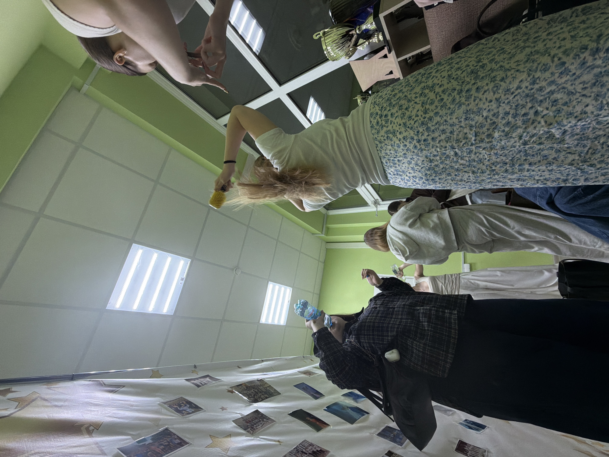The image shows an interior space, likely a classroom or event hall, in Perm, Russia. A group of people stand around a table. The table is draped with a white cloth adorned with photos and star patterns. A person in a plaid shirt and black pants is holding a plastic bottle filled with blue liquid. Another person is brushing their hair with a yellow brush. Above, fluorescent ceiling lights illuminate the room, which has green walls. Other individuals stand nearby, observing. A trophy sits on a shelf in the background.