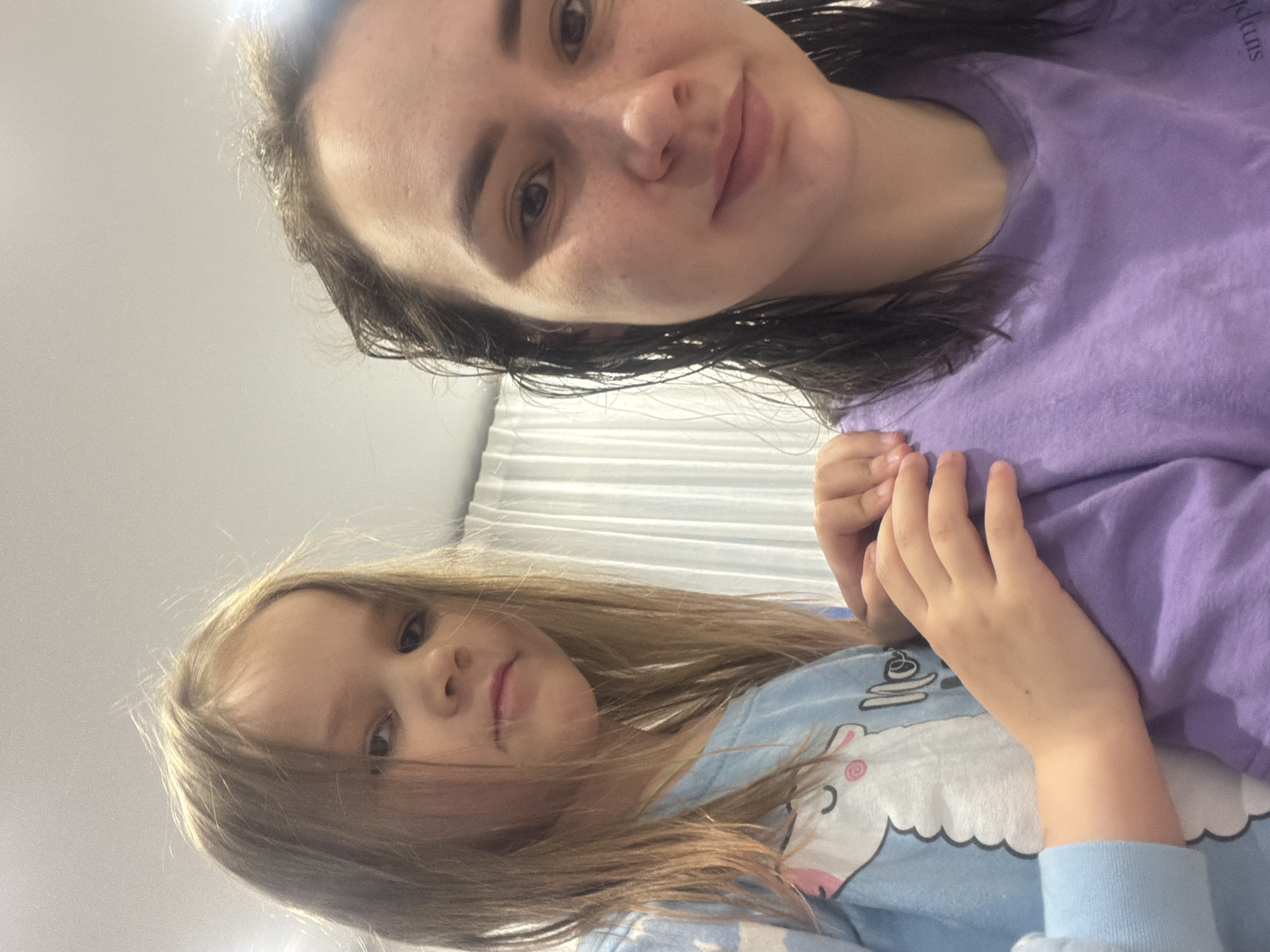 An image captured at an angle shows an adult and a young child, likely a mother and daughter, indoors. The camera appears to be looking down on them.
The adult, positioned towards the top right of the frame, has dark, possibly wet hair, pulled back from her face. She has fair skin, a light scattering of freckles, and dark eyes, looking directly at the camera with a soft, slight smile. She is wearing a light purple long-sleeved t-shirt. Faint white text is visible on her shirt, partially obscured, which appears to include "pluns" or "pkins" and part of an "S" towards the top right. Her left hand is visible, resting gently on the child.
The child, positioned towards the bottom left, has long, straight light brown or blonde hair. She has fair skin and dark eyes, also looking at the camera with a neutral, calm expression. She is wearing a light blue long-sleeved top featuring a white cartoon character, possibly a unicorn or llama, with pink details on its face and a blue mane, standing on a cloud. Her hands are near her chest, with her left hand resting on the adult's arm.
The background is a bright, plain light color, possibly a wall or a curtain, suggesting an indoor setting with even lighting. There are no strong shadows, indicating soft or diffused light. The overall mood of the image is calm and familial.