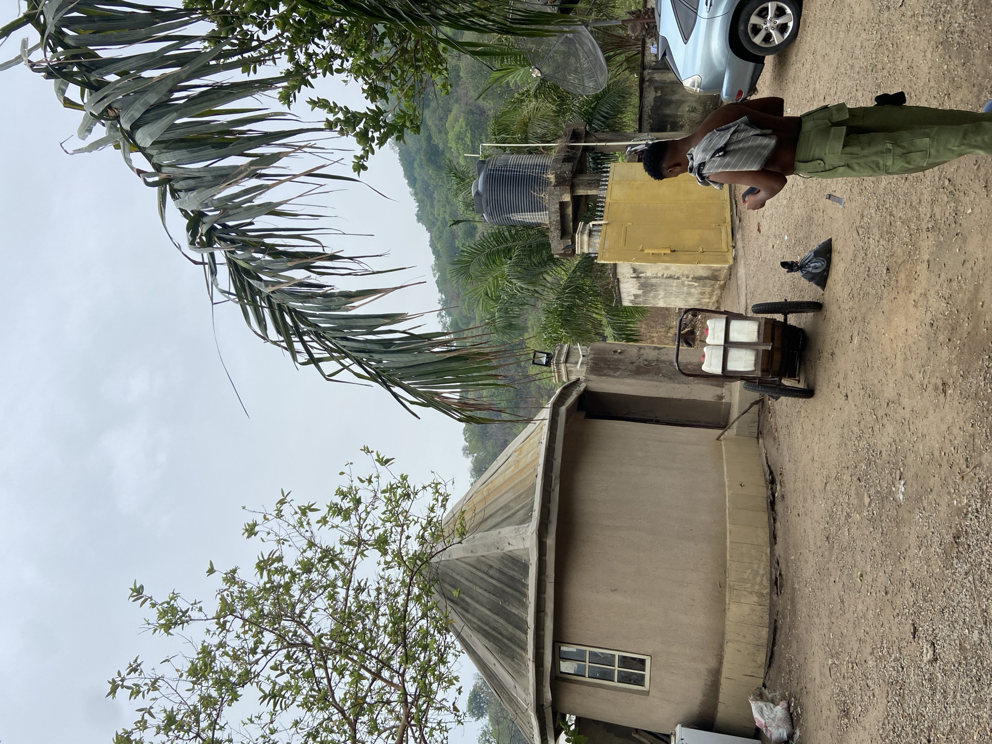The image shows a scene in Mararaba, Nigeria. A man stands near a building with a corrugated metal roof. He is wearing green pants, no shirt, and a striped cloth draped over his shoulder. Next to him is a two-wheeled cart loaded with several white containers. Behind the man, there's a light blue car and a yellow structure. In the background, a hillside with lush green vegetation can be seen, along with a black water tank and a satellite dish. The foreground is a dirt ground. A palm tree branch hangs in the top left corner of the image, and another tree is visible in the bottom left. The sky is overcast.