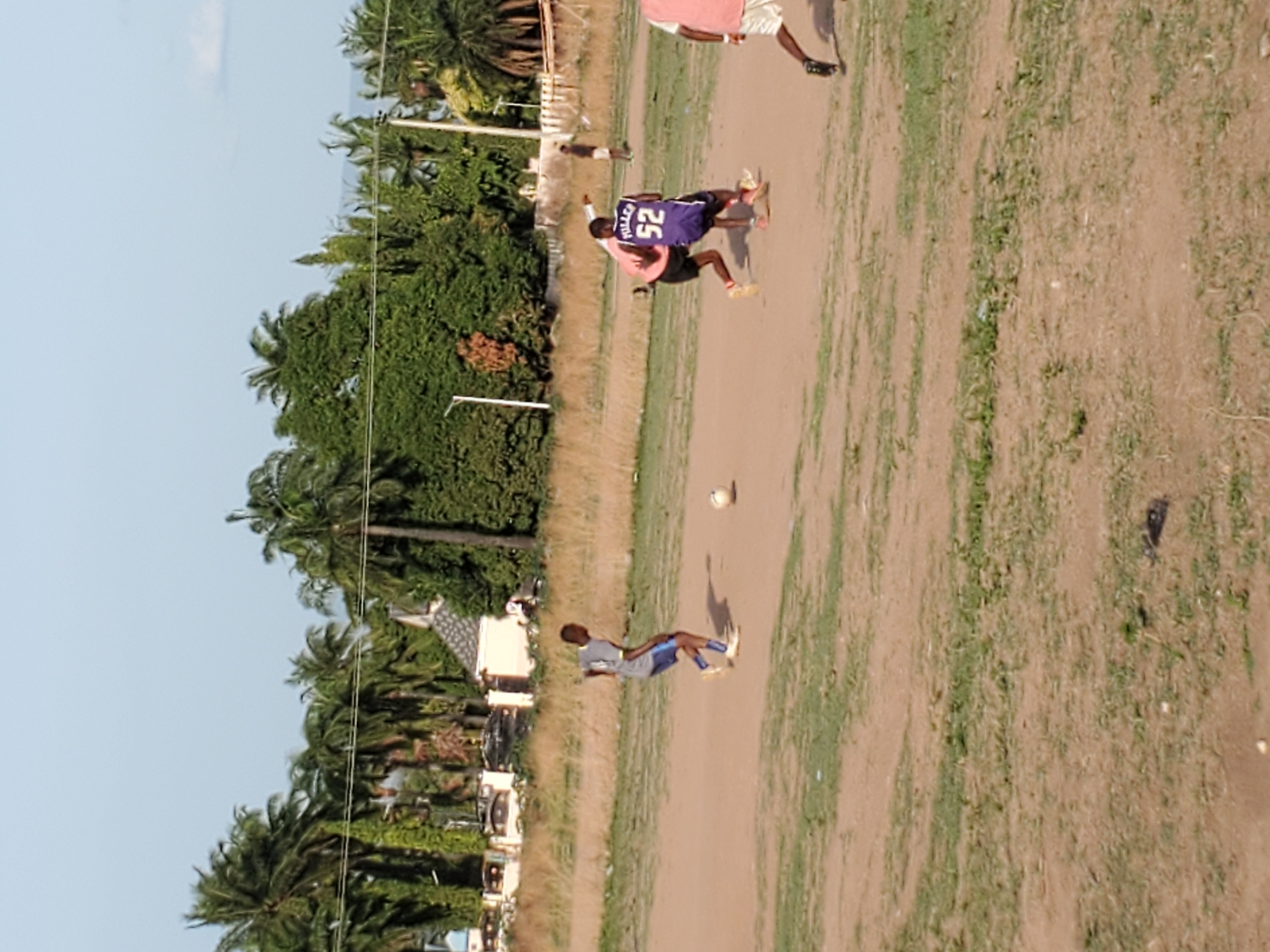 This image, captured in Mararaba, Nigeria, depicts an outdoor soccer (football) game in progress on a sunlit, unpaved field. The time of day is bright daylight, indicated by strong illumination and a clear, light blue sky with minimal cloud cover.
Several young men are the primary subjects. One prominent player, seen from the back, wears a purple jersey with the name "FULLER" and the number "52" clearly printed on it, paired with black shorts and bright yellow footwear. Another player nearby wears a light pink t-shirt and dark shorts. To the left, a player in a grey shirt and blue shorts stands, looking towards the ball. A white soccer ball rests on the dusty ground near this player. Another individual, partially visible in the upper right, wears a pink shirt and white shorts.
The playing surface is a mix of bare brown earth and sporadic patches of green grass, typical of an informal pitch. The setting is framed by a dense line of lush green trees, predominantly palm trees, stretching across the background. Several utility poles or posts are visible among the foliage, and some buildings can be faintly seen in the distance behind the trees, suggesting a semi-urban or rural environment. The scene conveys a sense of casual, recreational activity.