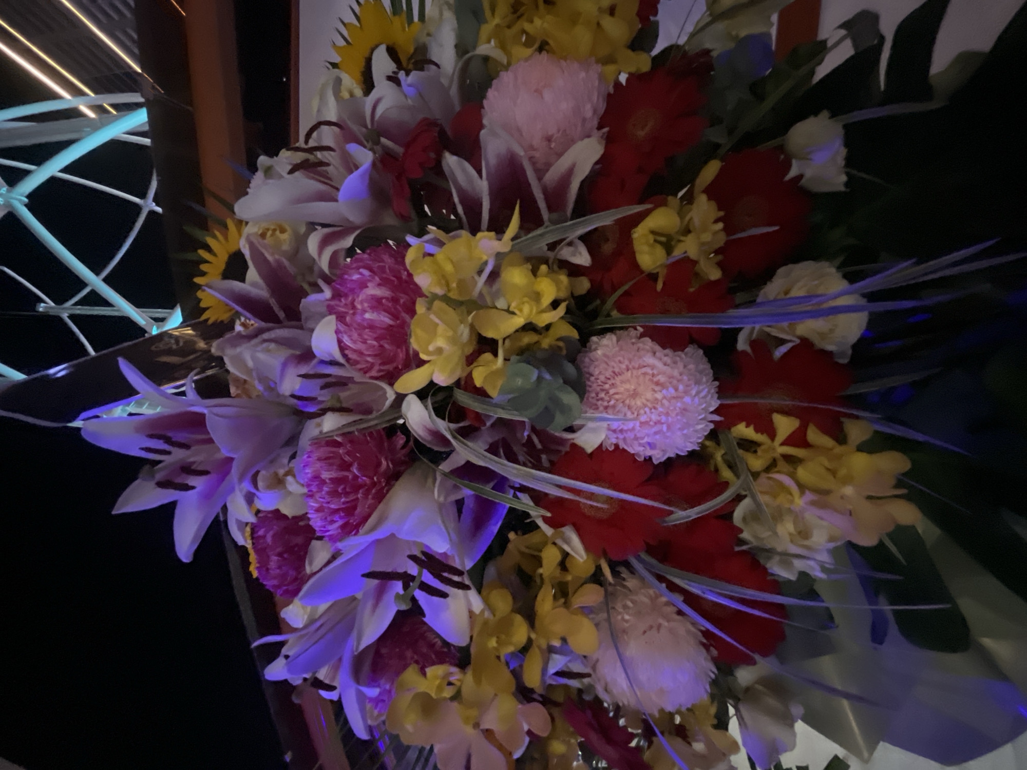 A large, colorful floral arrangement is displayed outdoors at night, illuminated by artificial lighting which casts a purplish hue on some of the flowers. The bouquet contains a variety of flowers including lilies, sunflowers, chrysanthemums, gerbera daisies, and orchids in shades of white, pink, red, yellow, and purple. The background features dark, indistinct shapes, with a partially visible structure that appears to be a Ferris wheel with illuminated supports, suggesting a location at a theme park or amusement area. The overall scene is likely a decorative display in an entertainment venue in the city of Da Nang, Vietnam, possibly near the Han River or an area with tourist attractions. There are no people or visible animals in the image.