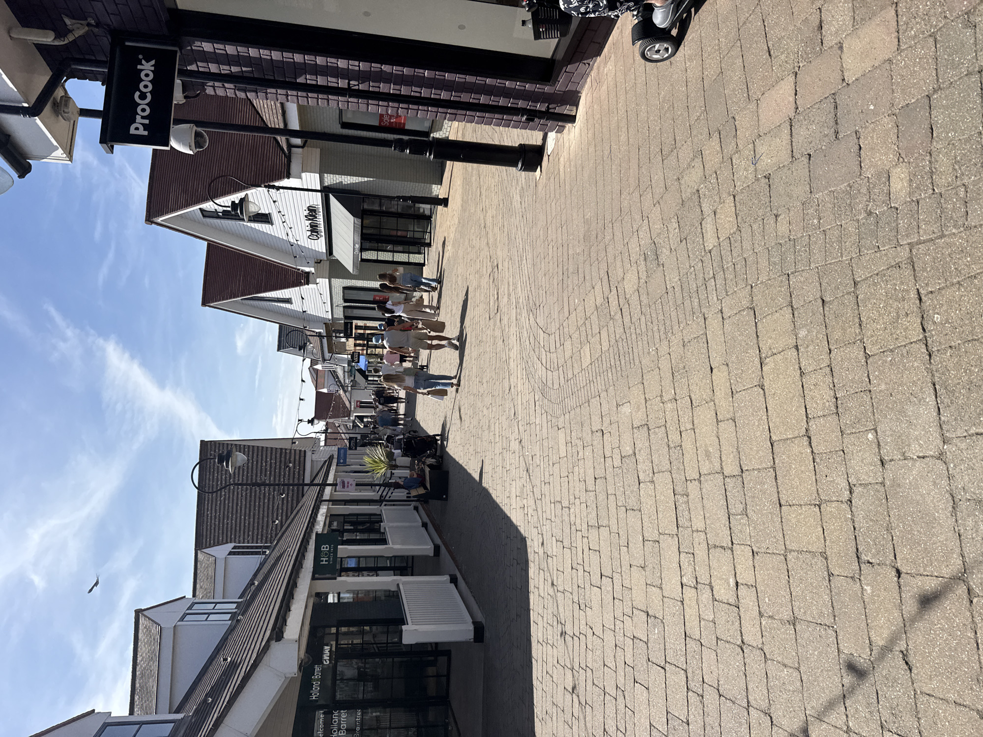 The image shows a sunny day at a retail outlet located in Braintree, United Kingdom. The paved walkway is lined with various stores, including Holland & Barrett, H&B, Columbia, and Calvin Klein. Several people are walking along the path, some are shopping, and one person is using a mobility scooter. The architecture of the buildings features white facades, brown tiled roofs, and prominent signage for the retail establishments. The sky is blue with scattered white clouds, and a bird can be seen flying in the distance. The ground is paved with rectangular bricks.