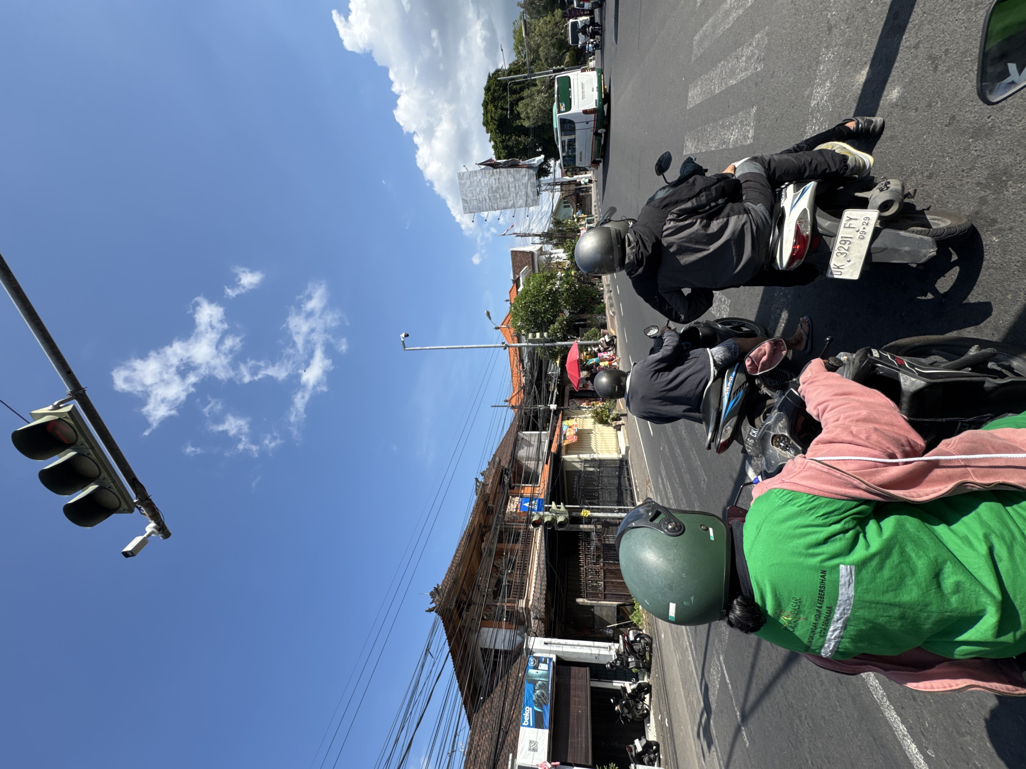 The image depicts a street scene in Denpasar, Indonesia, on a clear day with blue skies and scattered clouds. In the foreground, two individuals are riding motorcycles. The person in the closest motorcycle is wearing a green helmet and a vibrant green jacket with white text that reads "DENPASAR LINGKUNGAN HIDUP & KEBERSIHAN KOTA DENPASAR," indicating they are likely part of the Denpasar environmental and cleanliness department. They are also wearing a pink jacket. The second motorcyclist, in front of them, wears a black jacket and a gray helmet.
The street is paved with asphalt and marked with white pedestrian crossing lines. On the left side of the road, traditional Balinese-style buildings with intricate wooden carvings and tiled roofs are visible. A store with a "beko" logo is present. Traffic lights are also visible, with the red light illuminated, suggesting traffic is stopped. Several other motorcycles are parked on the side of the road.
Further down the street, more buildings with similar architectural styles line the road. A white bus with green accents is driving in the distance. A large, blank advertising billboard stands on the right side of the road. The overall environment suggests a busy urban setting in Denpasar.