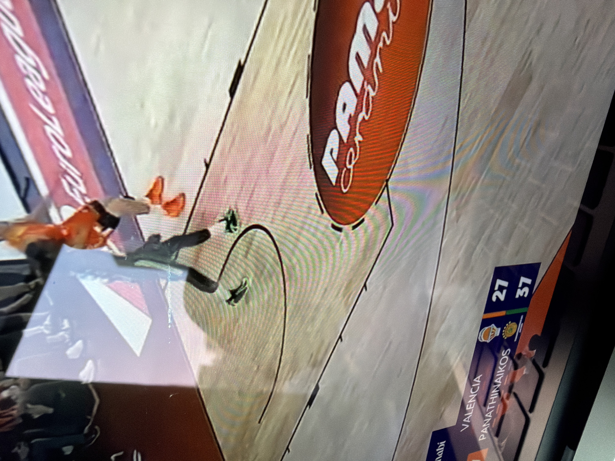 The image is a photograph of a screen displaying an ongoing indoor basketball game. The court features distinct white lines and a large red oval logo with white text spelling "Panini" at the center of the free-throw line area. Along the side of the court, advertising boards are visible, one displaying "EuroLeague" text on a blue and red background.
Two basketball players are discernible on the court. One player, dressed in an orange jersey, is positioned near the baseline, appearing to be on the ground or in a dynamic motion. Another player in dark attire stands nearby.
In the lower right corner of the screen, a digital score overlay shows the current game statistics. "VALENCIA" has a score of "27" points, accompanied by a team logo depicting a basketball in a hoop. "PANATHINAIKOS" has a score of "37" points, with a sun/leaf-like logo. Partially visible text below Valencia's score reads "dabi", likely part of "Abu Dhabi".
The photograph, captured in New York, USA, includes noticeable reflections on the screen's surface, which obscure parts of the displayed image but also reveal faint outlines of the surrounding environment where the photo was taken.