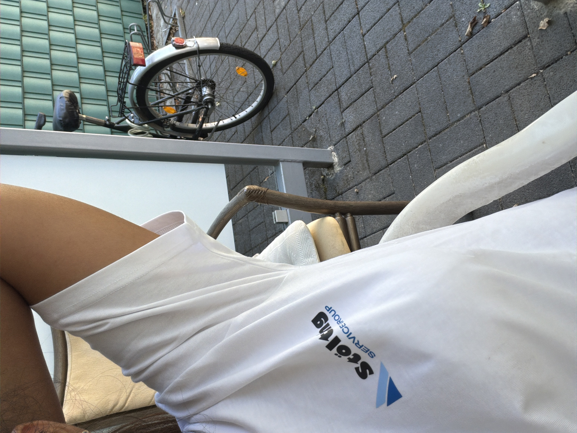 The image shows a person wearing a white t-shirt with a logo that reads "Stölting SERVICEGROUP". The logo is blue and black. The person is sitting on a chair, and a bicycle is visible in the background. The scene appears to be outdoors, with a paved area in front of a green fence. The image is taken from a close-up perspective, focusing on the t-shirt and the person's arm.