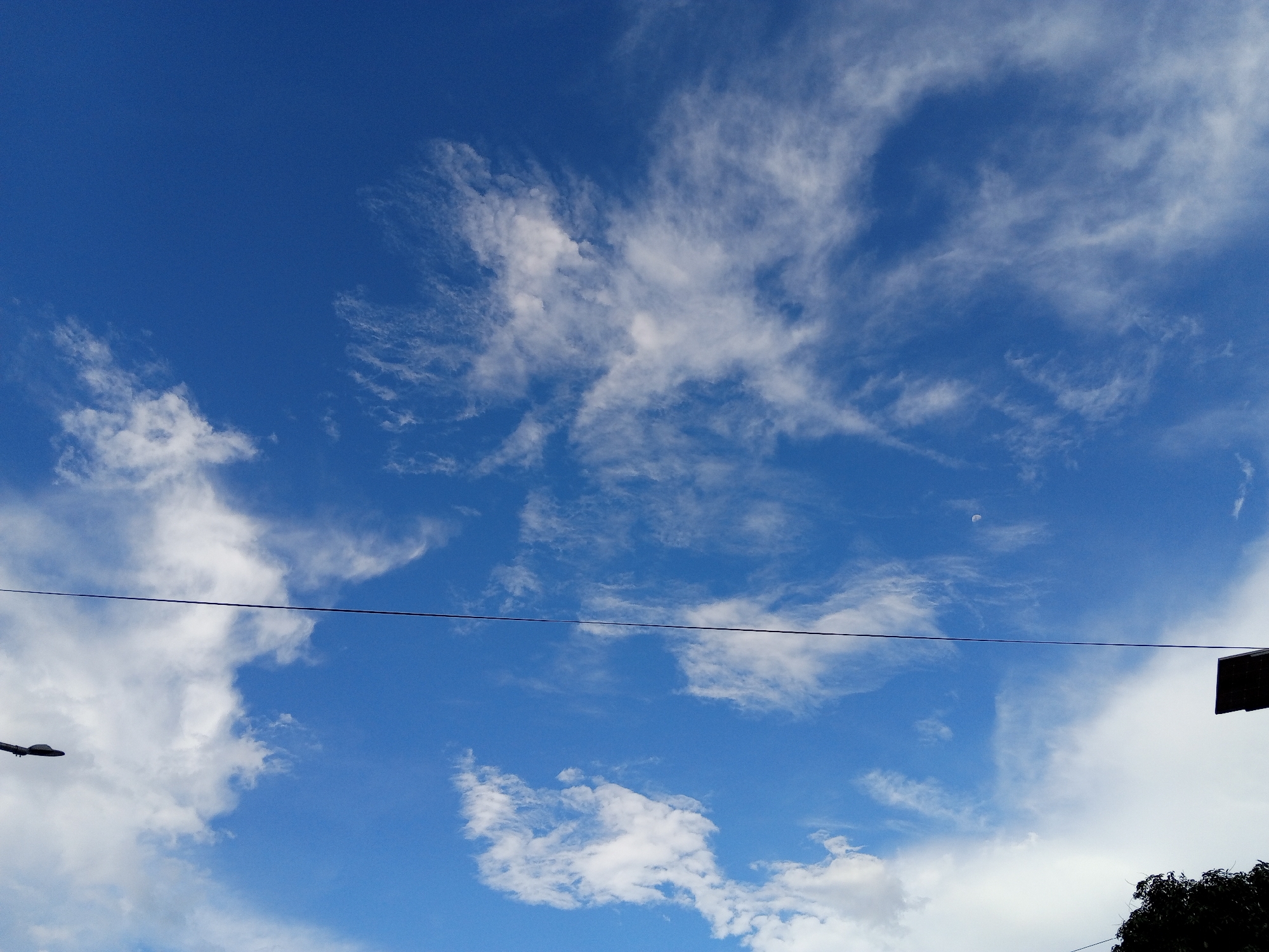The image depicts a daytime sky in Bireuen, Indonesia, characterized by a vibrant blue hue interspersed with various cloud formations. Wispy cirrus clouds are prominent in the upper portion of the frame, while cumulus clouds appear towards the bottom, some tinged with white. A single, thin wire runs horizontally across the center of the scene, possibly a power line. A street lamp is partially visible on the left. Vegetation, including a tree, is seen along the bottom right edge. A faint crescent moon can be seen in the top right portion of the frame.