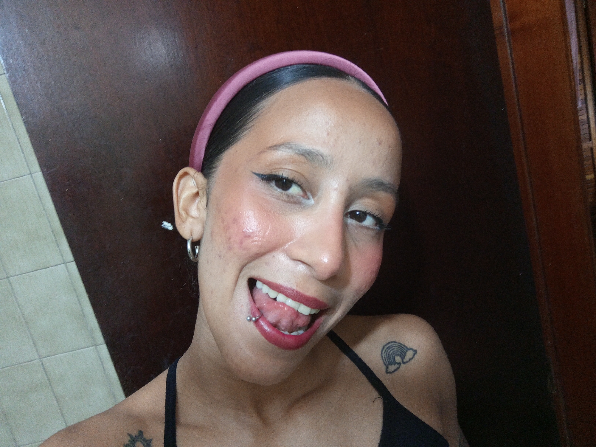 A person with a pink headband, visible skin imperfections, a tongue piercing, and shoulder tattoos poses for a selfie. The person is indoors, with a dark brown wall and a partially visible tile wall in the background. The city of Porlamar, Venezuela cannot be confirmed from the image.