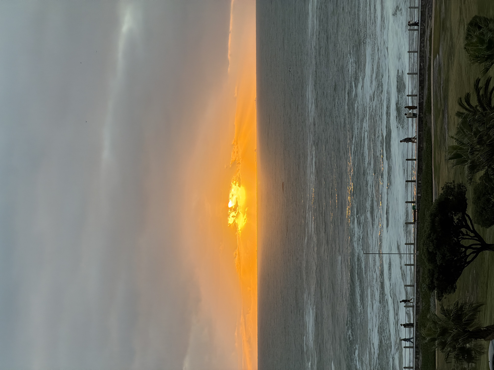 The image depicts a sunset over the ocean in Cape Town, South Africa, viewed from a promenade lined with trees and a concrete railing. People are walking along the promenade. The sunset appears as a bright orange glow through the clouds. The ocean is grey, with waves crashing against the railing.
