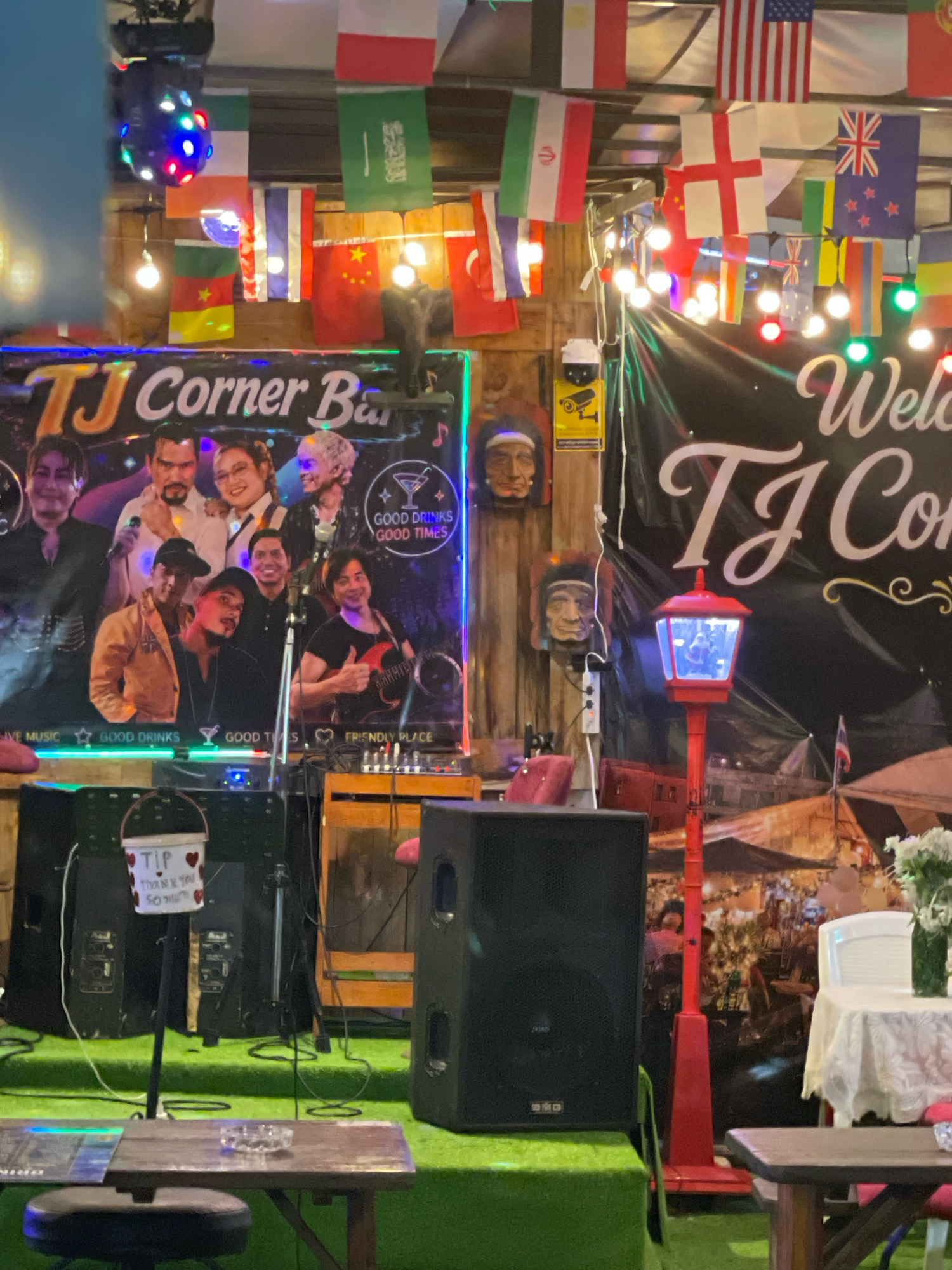 This image depicts the interior of TJ Corner Bar, located in Pattaya City, Thailand. The scene is filled with national flags hanging from the ceiling, creating a festive atmosphere. In the foreground, a stage area is set up with musical equipment, including speakers and a mixing board. A poster of the bar's band or performers is prominently displayed on the left side of the image. A red lamp post with a blue light and a bucket labeled "TIP" with hearts are also visible. The background features a mural or backdrop depicting a bustling street scene. The overall impression is that of a lively establishment that likely hosts live music performances.
