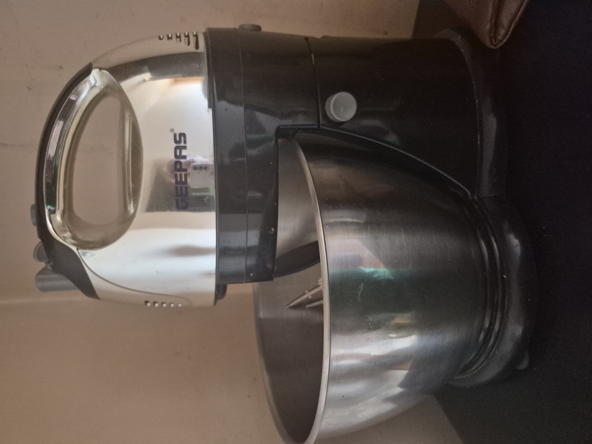 The media file displays a GEEPAS stand mixer, positioned on a surface in what appears to be an indoor setting in Yerevan, Armenia. The mixer's main body is black, featuring a metallic, silver-colored head with a prominent handle. The brand name "GEEPAS" is clearly visible, oriented vertically, on the side of the silver head. Several small, raised markings or ventilation holes are present on the top and side of the mixer head.
A large, polished stainless steel mixing bowl sits beneath the mixer head, housing a mixing attachment, likely a whisk or beater, though its exact form is not distinctly clear. The mixer head appears to be tilted back, indicating it might be in a raised position for bowl access, or the image is simply taken from a high angle. A round, light-colored button or control knob is visible on the black body of the mixer. The background is simple and indistinct, with a light wall or surface behind the mixer and a darker, soft-looking object (possibly furniture) to its right. No activity is taking place; the mixer is stationary and appears clean.