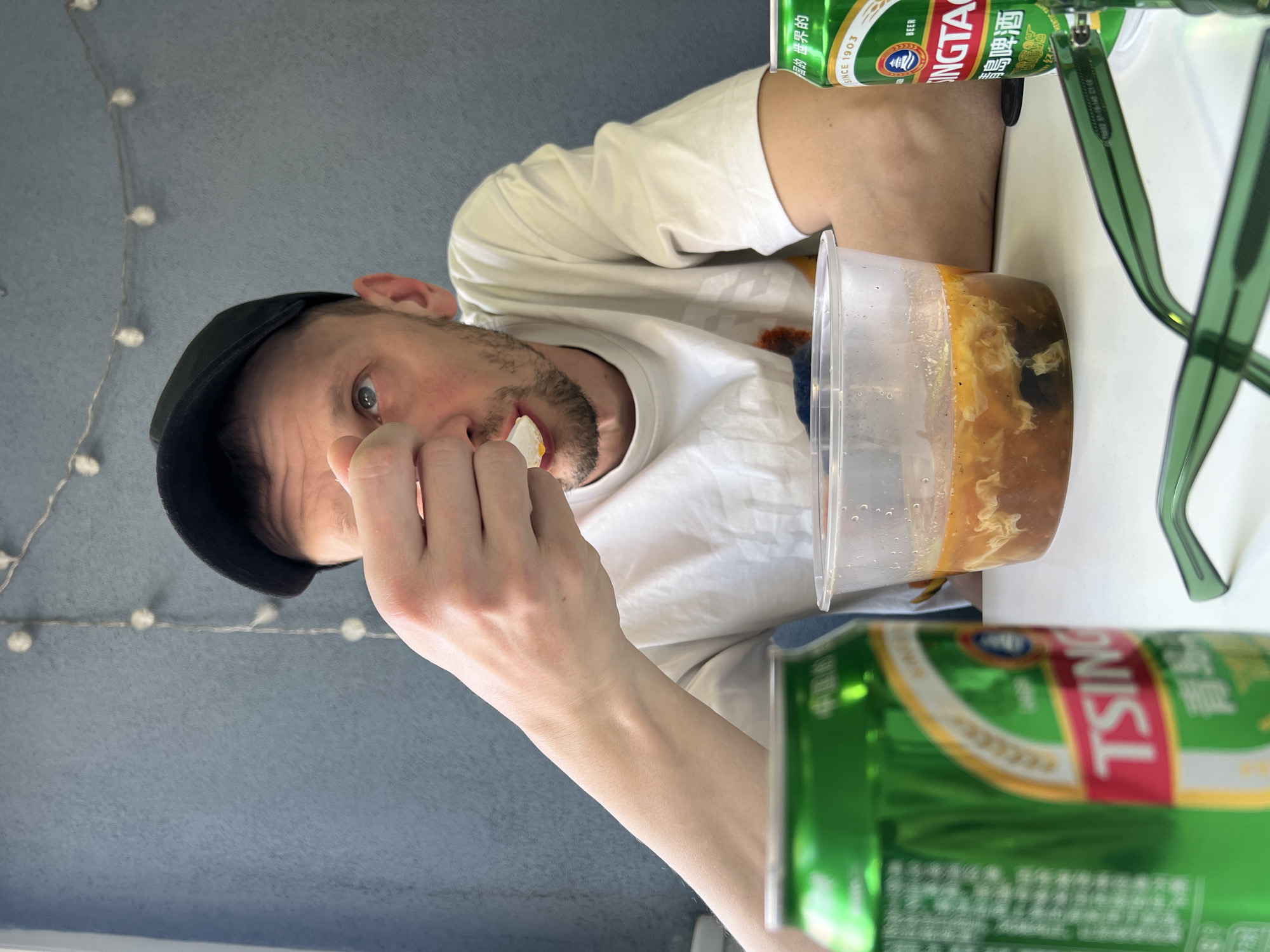 A fair-skinned man wearing a black cap and a white t-shirt is eating from a clear plastic container. He is holding a small piece of food with his fingers. Next to the container are two green cans of Tsingtao beer and a pair of green-rimmed glasses. The background is a gray-blue wall decorated with a string of lights. The location is likely indoors, but the city of Belgrade, Serbia, cannot be confirmed from the visual information provided.