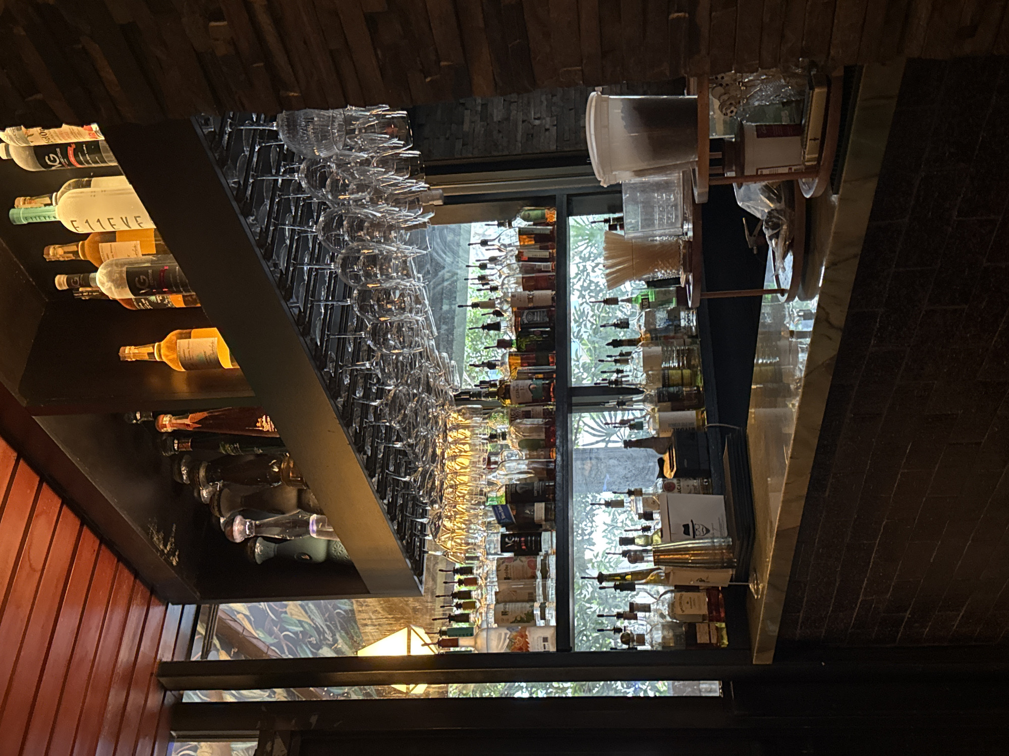 The image depicts the interior of a dimly lit bar. In the foreground, a bar counter made of marble sits atop a dark brick base. Behind the counter, shelves are stocked with an assortment of liquor bottles, glassware hanging from a rack above, and other bar accessories. The background shows a window with greenery visible, suggesting an outdoor view. The overall ambiance is that of a typical bar or restaurant setting. The location is identified as Fort Lauderdale, United States of America.