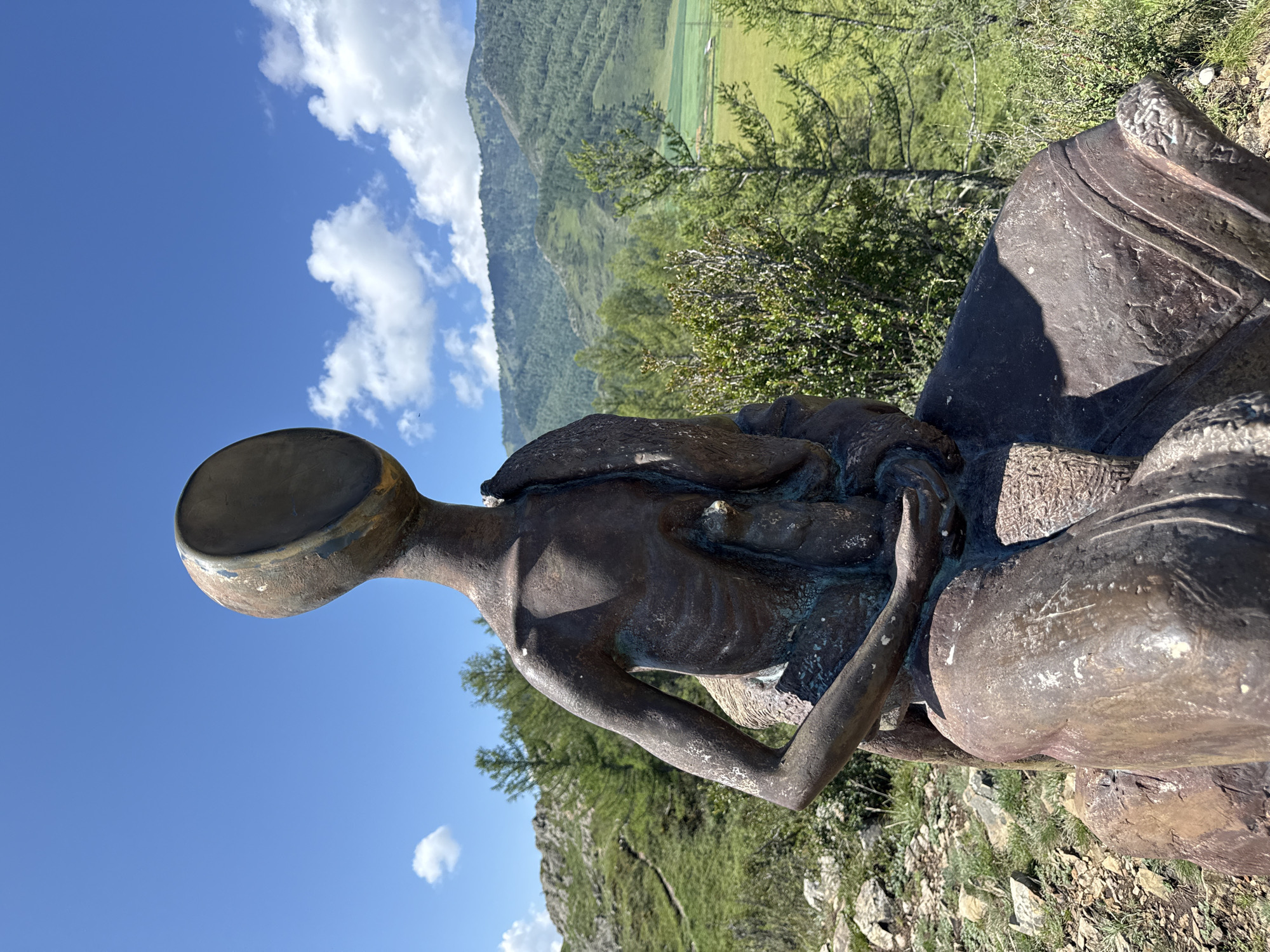 The image shows a bronze sculpture of a seated figure with an empty, bowl-like head. The sculpture is positioned outdoors, in a natural landscape. The figure is dark brown and appears to be aged, with visible texture and some green patina. It is seated on what seems to be a rough, stone base.
The background features a lush, green mountainous terrain under a bright blue sky with scattered white clouds. The foreground and middle ground show grassy slopes, rocky outcrops, and scattered evergreen trees. A winding path is visible on the left side of the image, leading up a hill. In the distance, there are more rolling hills and a field that appears to be cultivated, with some small buildings or structures.
The specific location is identified as Bichiktu-Boom, Russia. The sculpture is situated in a scenic, elevated spot offering panoramic views of the surrounding natural environment. The overall impression is one of a contemplative or symbolic artwork placed within a striking natural setting.