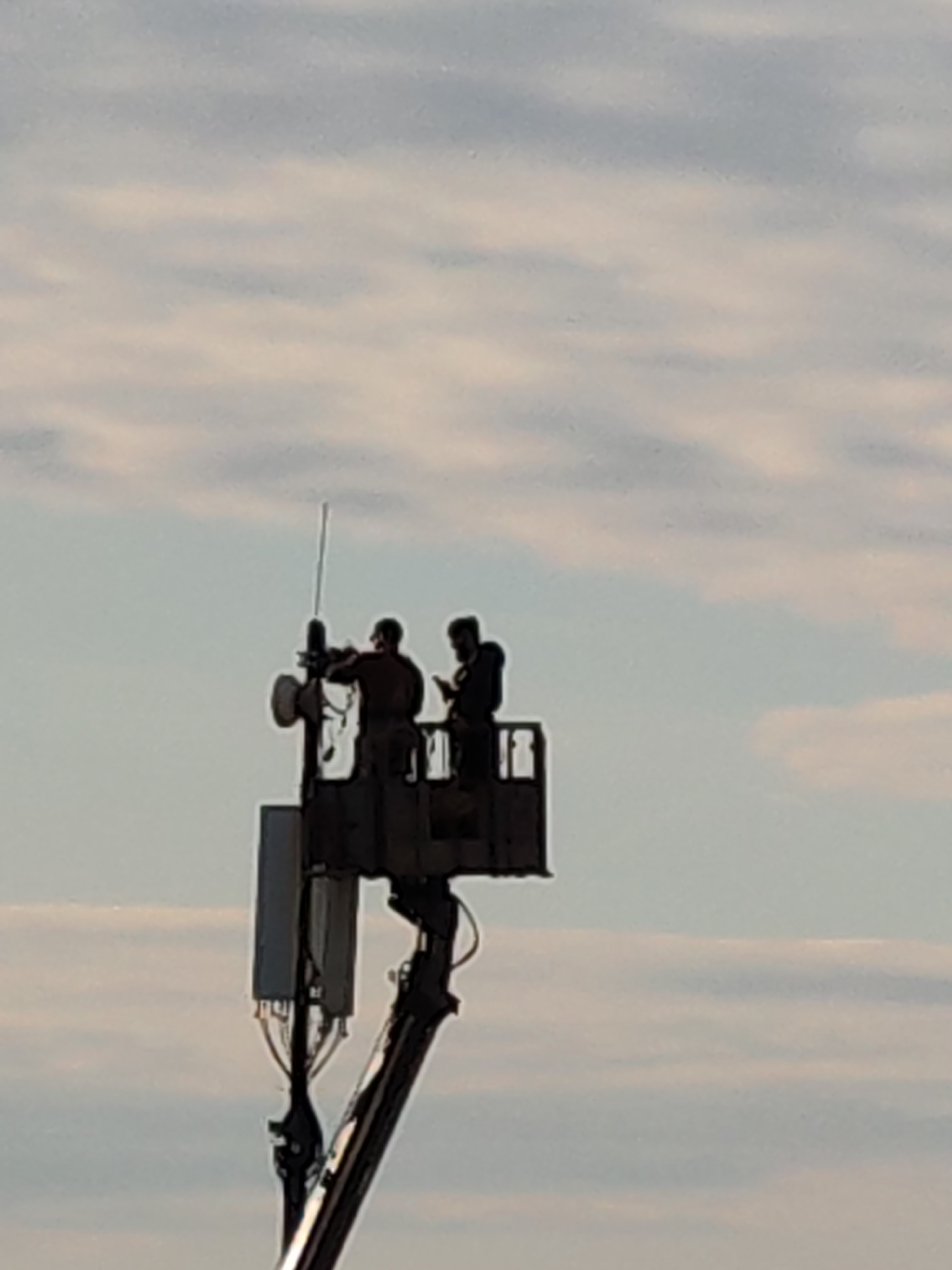 The image captures two individuals working on telecommunications equipment from an elevated lift platform, set against a cloudy sky. The scene is located in Podolsk, Russia.
The two subjects are silhouetted, indicating the sun is behind them, suggesting either early morning or late afternoon. They appear to be technicians or engineers, wearing work attire. One individual is positioned closer to a metal mast, which is fitted with a tall, slender antenna and several rectangular modules, likely components of a cellular base station or radio communications array. This person appears to be actively engaging with or adjusting the equipment. The second individual stands further back in the work basket, possibly holding a mobile device such as a smartphone or tablet, which could be used for monitoring, communication, or data input related to the task.
The lift platform itself is part of a hydraulic crane or boom lift, extended high into the air to reach the telecommunications infrastructure. Cables are visible running from the equipment boxes downwards along the support structure. The sky is largely overcast with soft, streaky clouds, creating a diffused light that emphasizes the silhouettes of the workers and equipment. No specific text is visible on the equipment or the workers' clothing. The overall impression is one of focused technical work being performed at height in an outdoor environment.