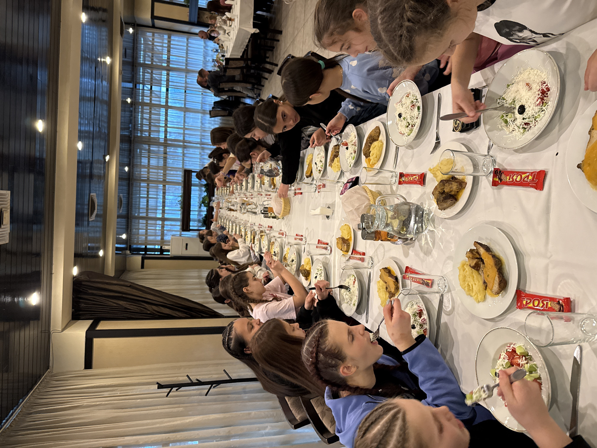 A long, white-clothed table is set for a meal in what appears to be a restaurant or banquet hall. The table stretches into the distance, lined with plates of salad, potatoes, and meat, interspersed with glasses and candy bars. A large group of children, mostly girls with braided hair, are seated along the table, eating. The location is Etropole, Bulgaria. Beyond the children, the background shows other tables with people seated and dining. Large windows allow natural light to illuminate the scene. The environment appears well-lit and spacious.