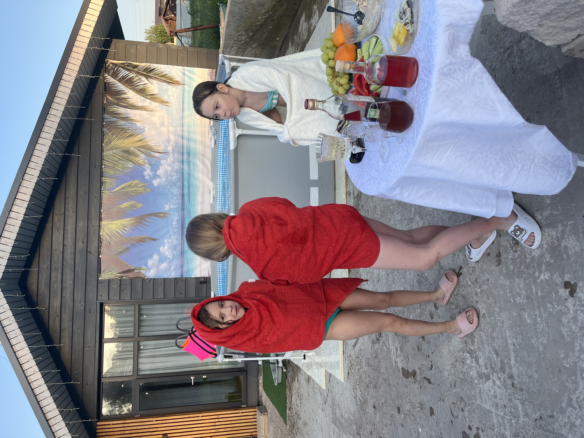 The image shows three girls standing near a pool in what appears to be the backyard of a house. Two of the girls are wrapped in red towels and wearing sandals. One girl is wearing pink sandals and the other is wearing white sandals. The third girl is wearing a white robe over a swimsuit. A table with a white tablecloth is present, with various fruits, drinks, and food items on it. Behind the girls, there is a house with a dark wood facade and a backdrop of a beach scene. The location is Новая Чернушка, Russia.