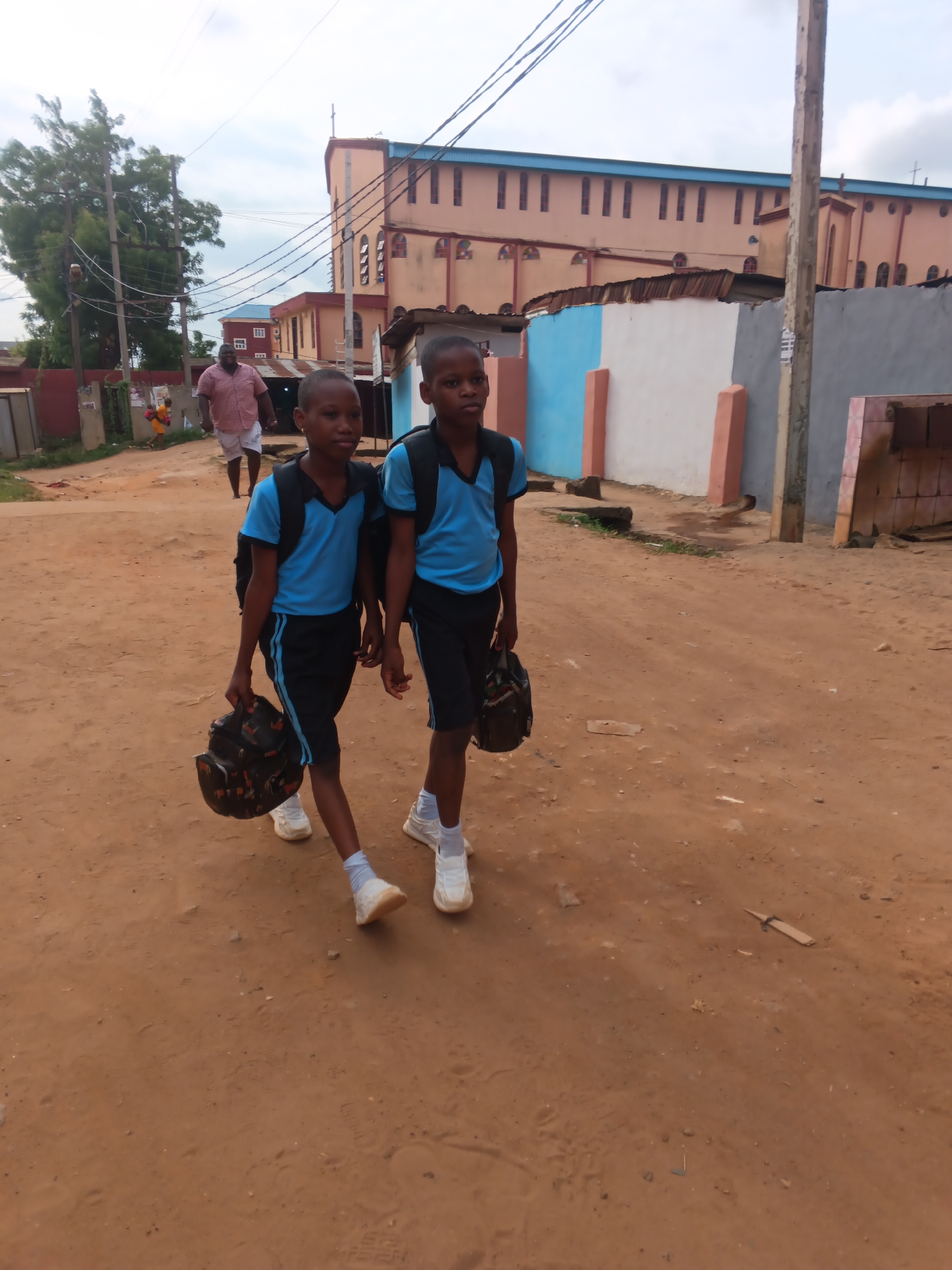 Two young boys in blue shirts and black shorts walk down a dirt road. Both boys wear white socks and white sneakers and have backpacks slung over their shoulders. They both carry a dark bag in their hands. In the background, an adult man walks in the same direction. Behind them, a building painted in shades of beige and pink can be seen. The location is Onitsha, Nigeria, as evidenced by the architectural styles, coloring and road.