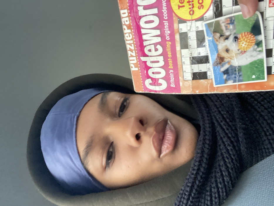 A person with dark skin and dark hair, covered by a blue satin durag and an olive-green hooded sweatshirt, is captured in a close-up, selfie-style image. A black knitted scarf is visible around their neck. The individual's lips are slightly pursed, and they gaze directly towards the camera. They are holding a pink and orange "PuzzliePag Codeword" puzzle book, partially obscuring the lower right side of their face and upper body. The cover displays "Britain's best-selling original codeword" text and features an image of a Jack Russell Terrier-type dog holding an orange spiky ball in its mouth, set against a green field and distant buildings. The background is an indistinct, light grey surface, providing no visual cues to determine a specific location within Paris, France, or any other geographic area.
