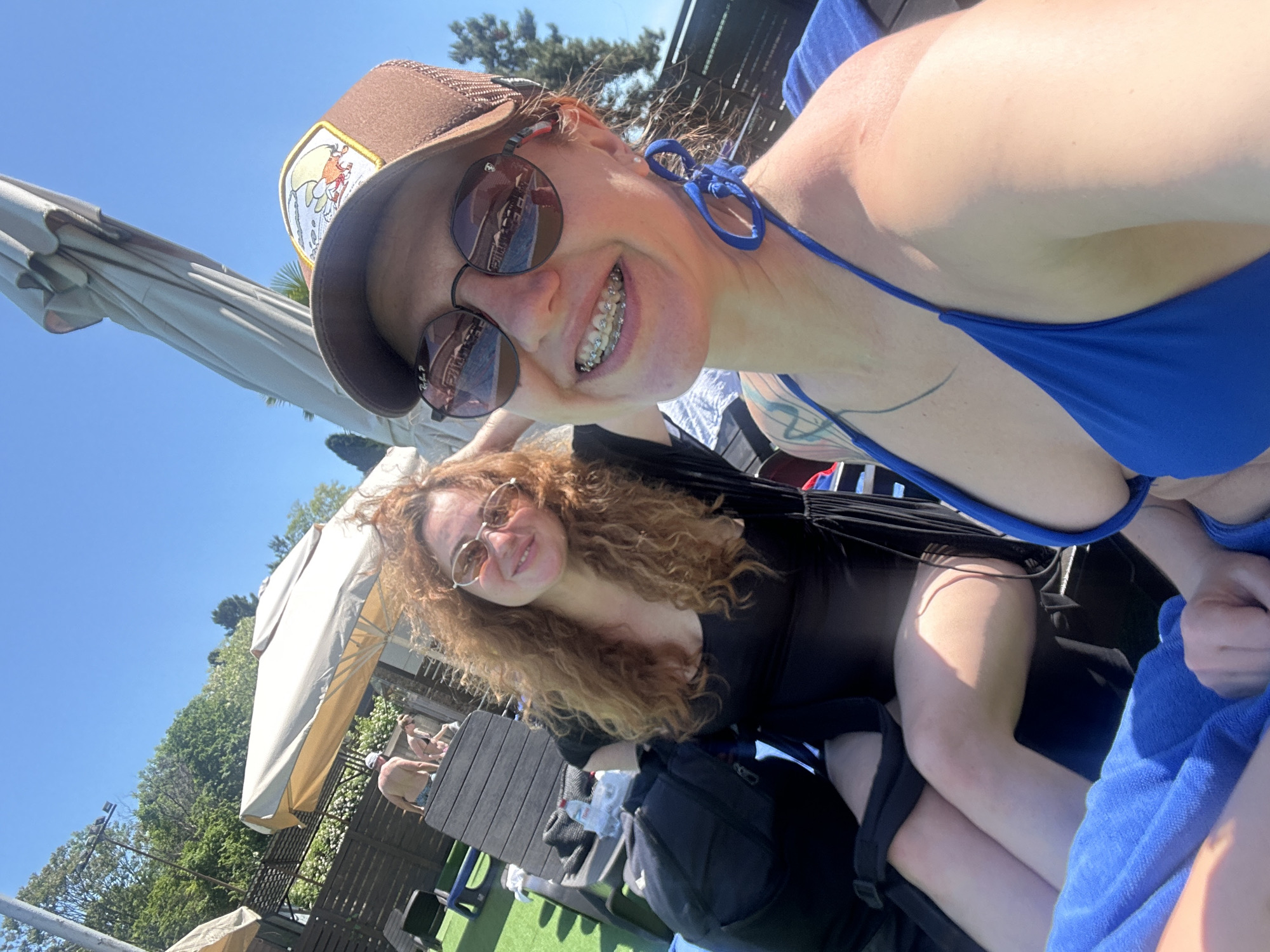 Two women are posing for a selfie on a sunny day. The woman in the foreground is wearing a brown trucker hat with a patch, round sunglasses, and a blue bikini top. She has red hair and braces. A tattoo is visible on her upper arm. The woman in the background has curly red hair and round sunglasses. She is wearing a black top. A black backpack is visible. They are sitting near a swimming pool, and a white umbrella and vegetation can be seen in the background. The image might be taken in Sochi, Russia.