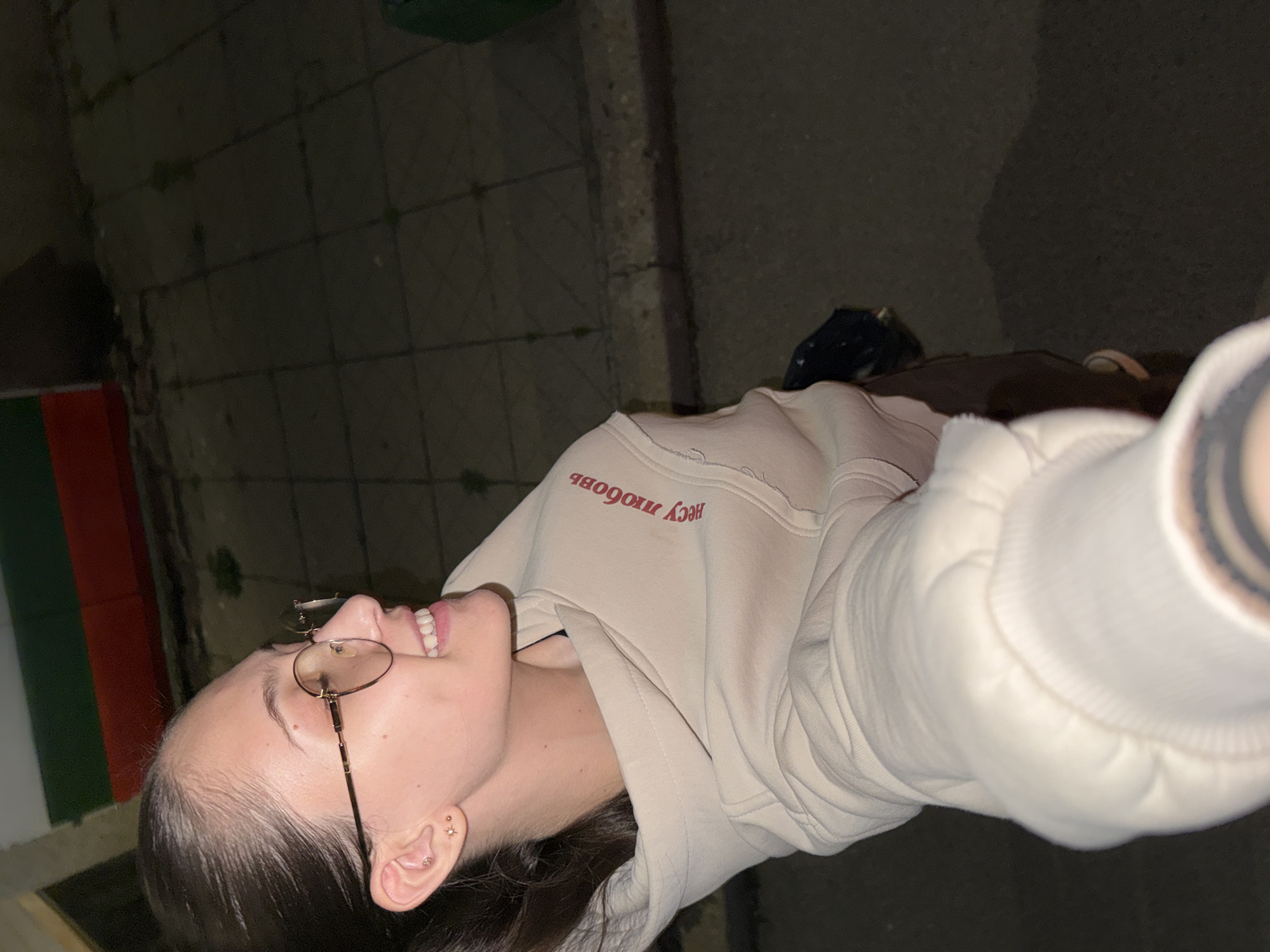 A selfie photo taken outdoors at night shows a young woman from the chest up, looking upward and smiling. She is wearing glasses with round frames and a light-colored hoodie with red text written in Cyrillic. The background shows a tiled ground and part of a wall painted in green, red, and white, possibly indicating a location in Sofia, Bulgaria, given the colors of the Bulgarian flag. The lighting suggests it is dark outside with artificial light sources nearby.