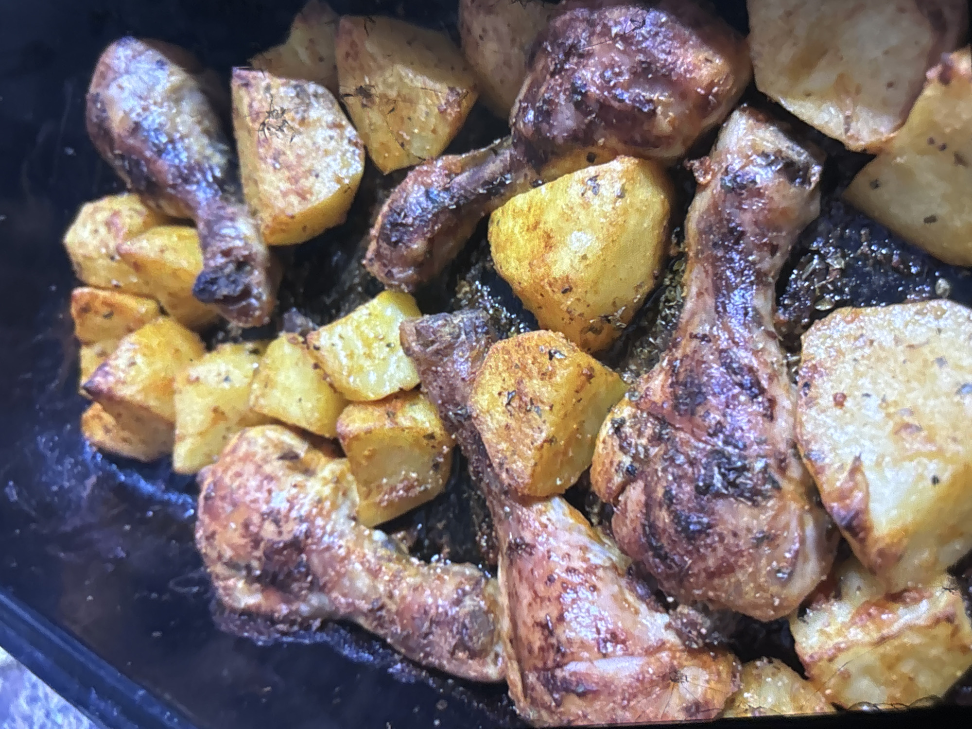 This is a close-up, top-down shot of roasted chicken drumsticks and potato wedges in a dark baking pan. The chicken pieces are golden brown with some darker, crisped skin. The potato wedges are also golden brown and appear seasoned with herbs. The dish is likely homemade and presented in a rustic manner. There are no visible people or any indications of a specific location in Orihuela, Spain. The focus is entirely on the food.