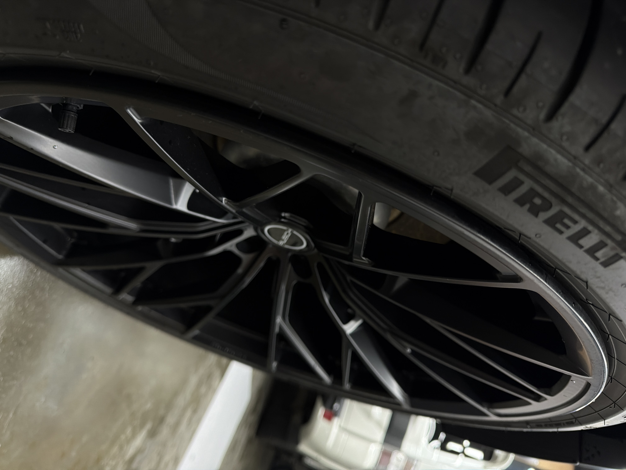The image shows a close-up of a car wheel and tire. The tire is black and has the word "IRELLI" visible on the sidewall. The wheel is also black, with a multi-spoke design. A car is visible in the background, out of focus. The environment is a garage. There is no evidence to suggest that the image was taken in Krasnoyarsk, Russia.