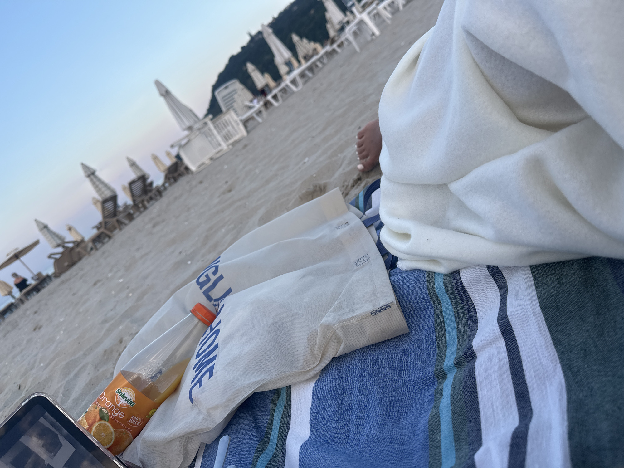 The image depicts a sandy beach with numerous lounge chairs and umbrellas in the background, suggesting a public beach area. The foreground shows a blue and white striped towel on the sand, with a white fabric bag and a plastic bottle of "Solevita Orange 100% Juice" lying on it. A person's foot, covered by a white blanket, is visible on the right side of the image. The scene appears to be set at Golden Sands beach, a popular resort located north of Varna, Bulgaria, known for its wide sandy shores and abundance of amenities for tourists. The sky is clear with a light blue hue, indicative of daytime.