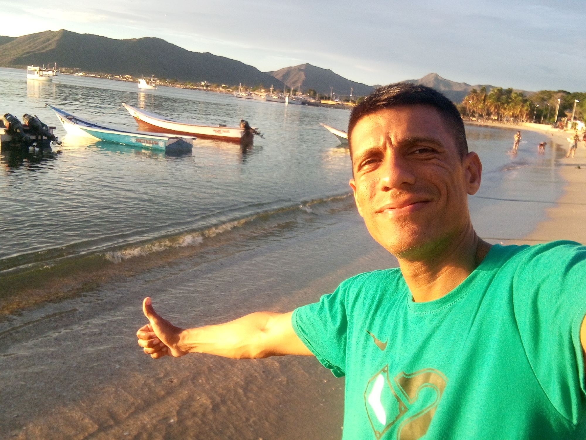 In this outdoor, daytime selfie, a man with short, dark hair and tanned skin stands on a beach, extending his right arm to give a thumbs-up. He is wearing a bright green t-shirt with a dark graphic design on the front. The background features a calm body of water with several boats anchored. Two of the boats are more prominent in the foreground: one is a narrow, blue and white vessel with two outboard motors attached, and the other is a longer, white boat with a reddish stripe. Further in the distance, more boats and a shoreline with buildings and palm trees are visible. Beyond the shoreline, a mountain range with a dark green, rugged slope dominates the horizon under a bright, hazy sky. The man's pose and the setting suggest a leisurely vacation or a positive experience at this coastal location. The scene is indicative of a beach in Juan Griego, Venezuela, known for its picturesque bay and mountainous backdrop.