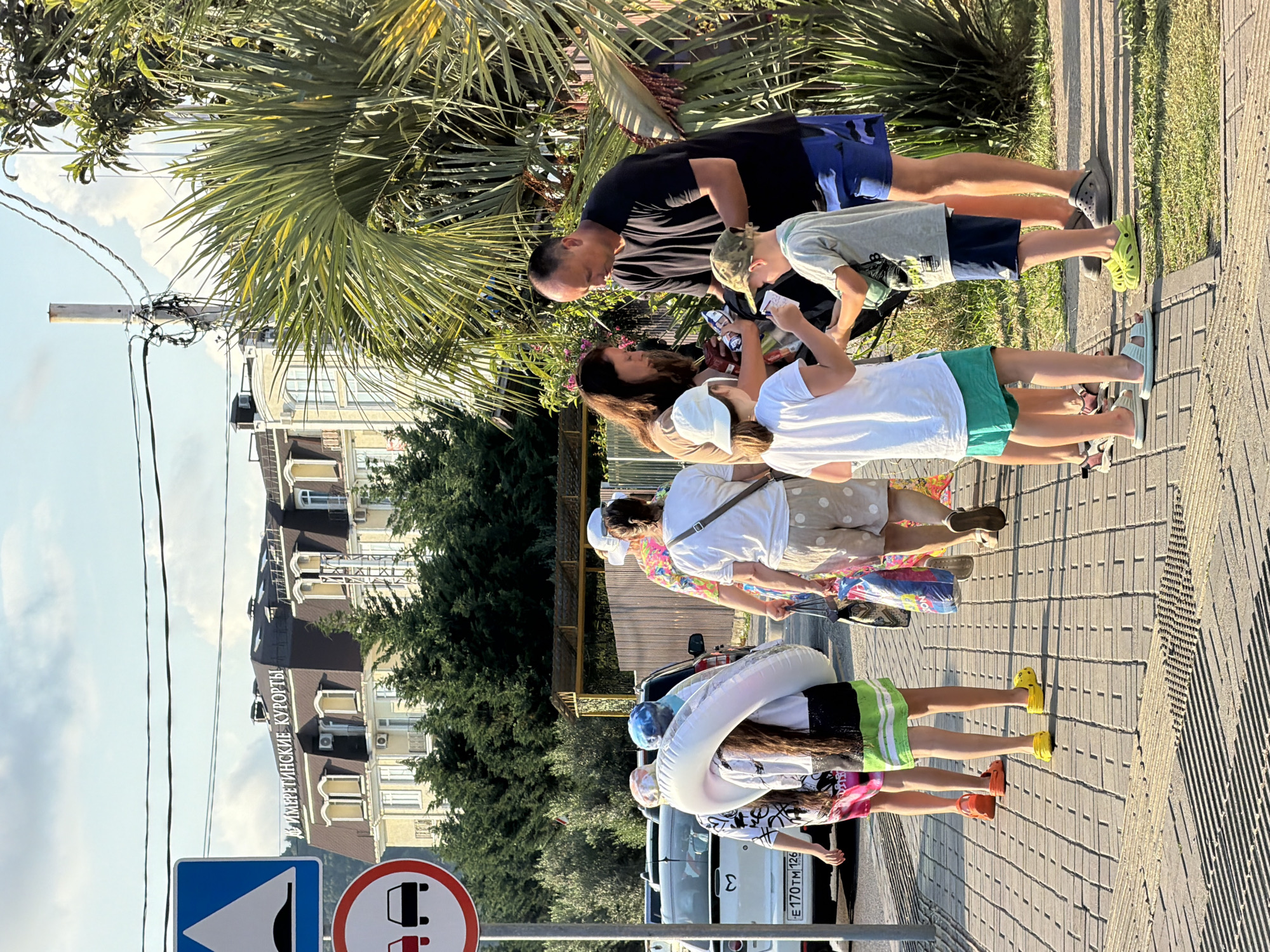 In the city of Sirius, Russia, on a sunny day, a group of people walks along a paved sidewalk lined with palm trees. In the background, a light-colored building with arched windows and a dark roof bears the inscription "ИМЕРЕТИНСКИЕ КУРОРТЫ" (Imeretinsky Resorts). A speed bump sign and a "no passing" sign are visible on the left. A man in a black t-shirt and blue shorts is interacting with children, handing something to a boy wearing a green cap and a gray t-shirt. Beside them, a woman with a white hat holds a bag, and another woman carries shopping bags. A young girl wearing a white shirt and green shorts is also present, along with another girl carrying a large inflatable ring. Cars are parked on the street behind them. The scene suggests a leisurely stroll, possibly towards a beach or resort area.