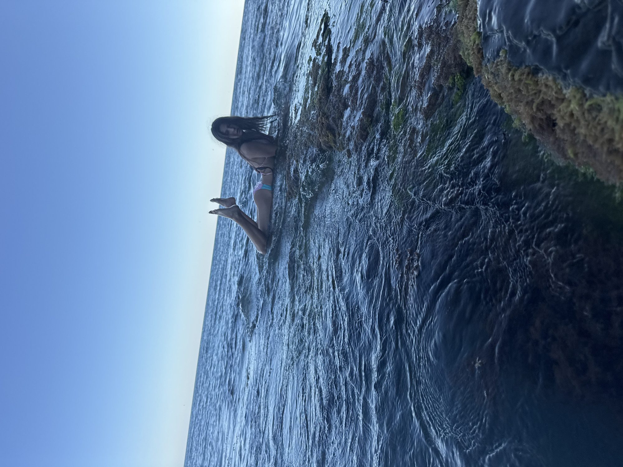 In the coastal waters of Dyurso, Russia, a young woman lies prone on a submerged rock formation. The clear blue sky forms a stark backdrop to the scene. Her body is partially submerged in the calm, dark blue sea, with her legs extended upwards. The rock she rests on is covered in algae and other marine growth, adding a textured, natural element to the scene. The gentle waves create ripples across the water's surface, suggesting a peaceful and tranquil atmosphere. The woman's gaze is directed towards the camera, her long, dark hair spread out around her head.