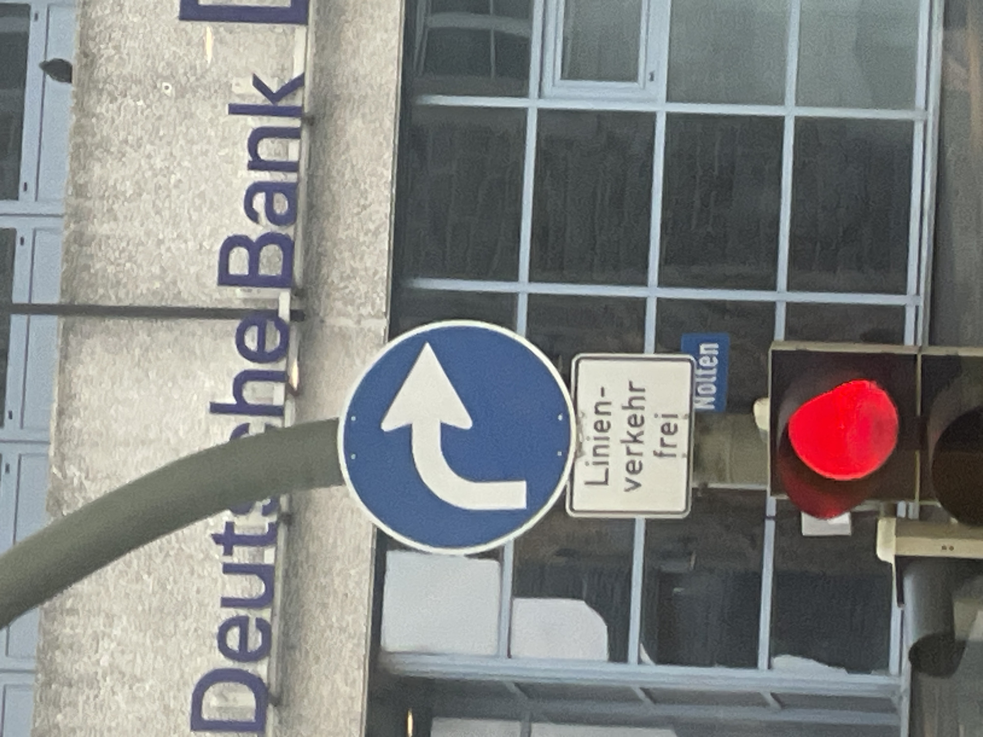The image shows a street scene in front of a Deutsche Bank building. A traffic light displays a red signal. Above the traffic light, there are two traffic signs. The upper sign is a blue circular sign with a white arrow indicating a right turn. Below this, a white rectangular sign reads "Linien- verkehr frei" which translates to "Public transport free". Underneath this sign, a smaller blue sign has white text that appears to say "Nolten". The building has large windows and the facade is textured. The lighting suggests it is daytime, possibly overcast.
