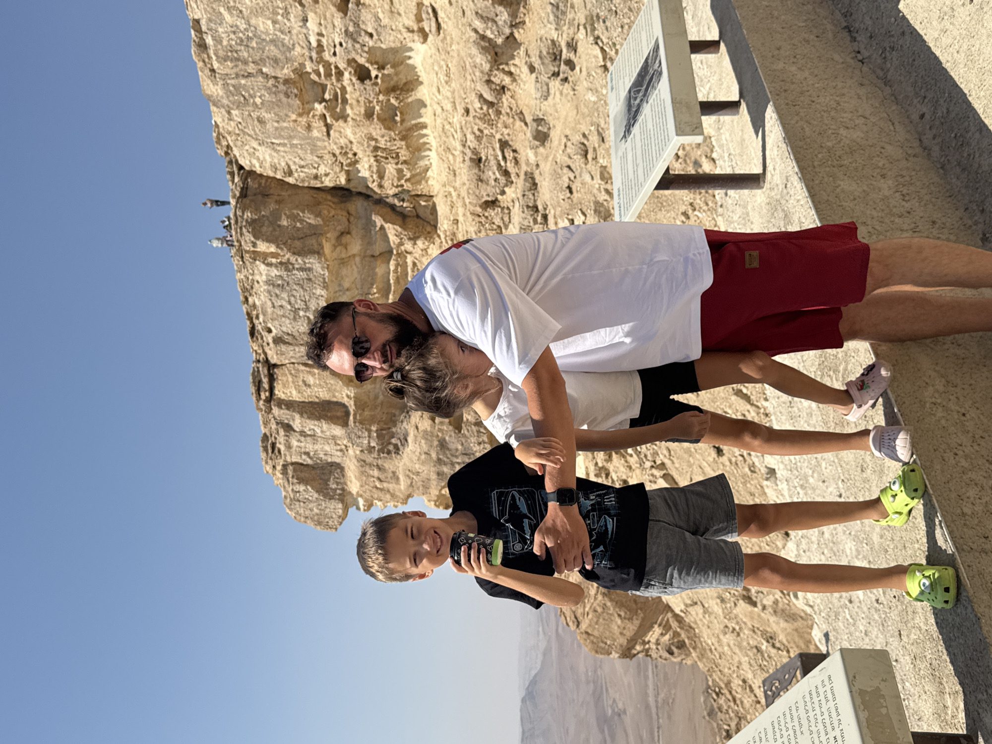 In Mitzpe Ramon, Israel, a man in a white t-shirt and red shorts embraces a young girl. To his left, a young boy in a black t-shirt and gray shorts holds a cup. They stand on a stone walkway overlooking a desert landscape with a large rock formation behind them. Several other figures are visible on top of the rock formation. A metal information stand is positioned to the right of the group. The sky is clear and blue.